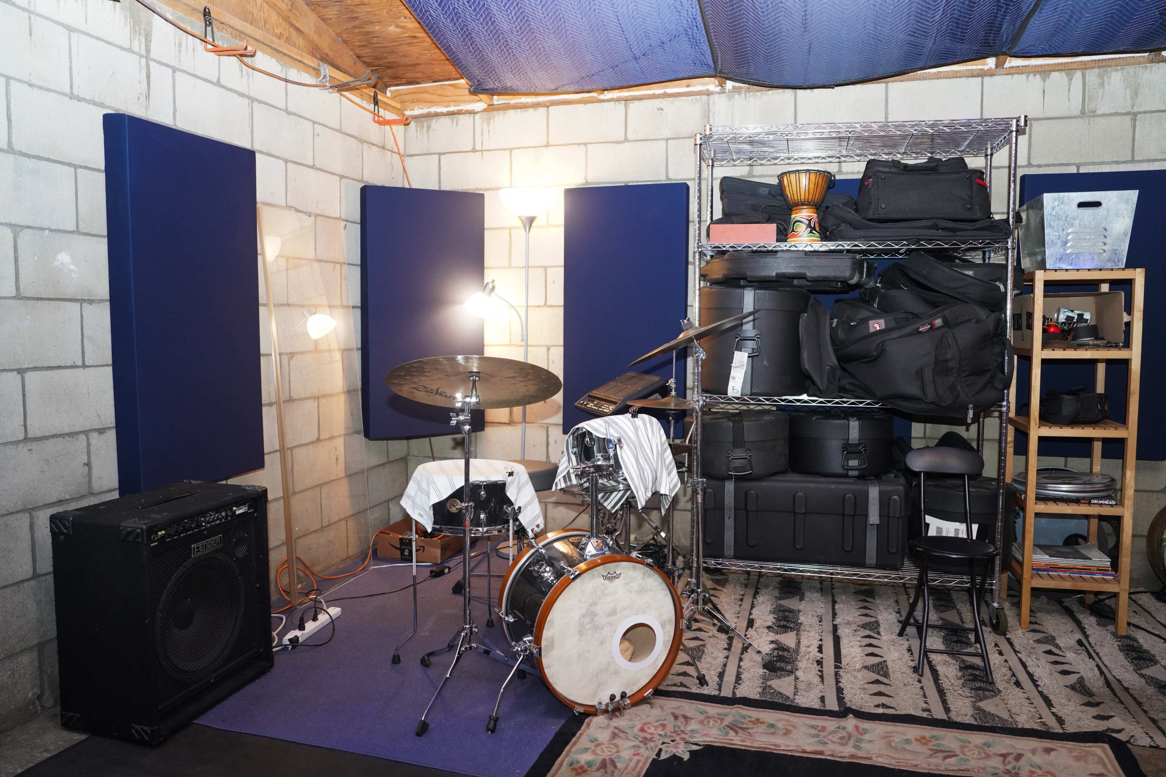 The image depicts a small music practice room featuring a drum set, amplifiers, and various storage containers organized on shelves.