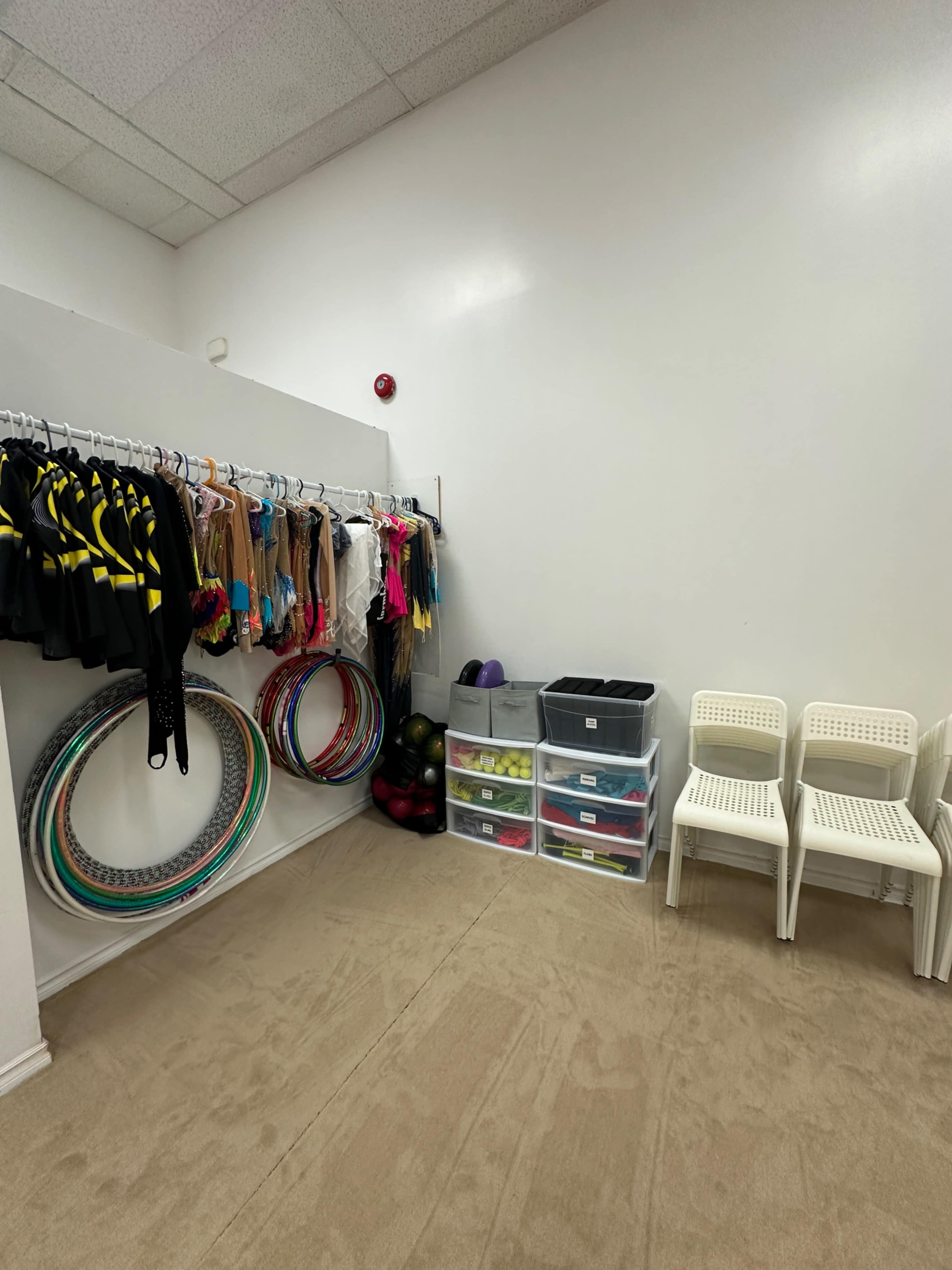 The image shows a small room with colorful costumes and accessories hanging on a rack, several hoops and balls arranged on the floor, plastic storage containers stacked, and two white chairs positioned against the wall.