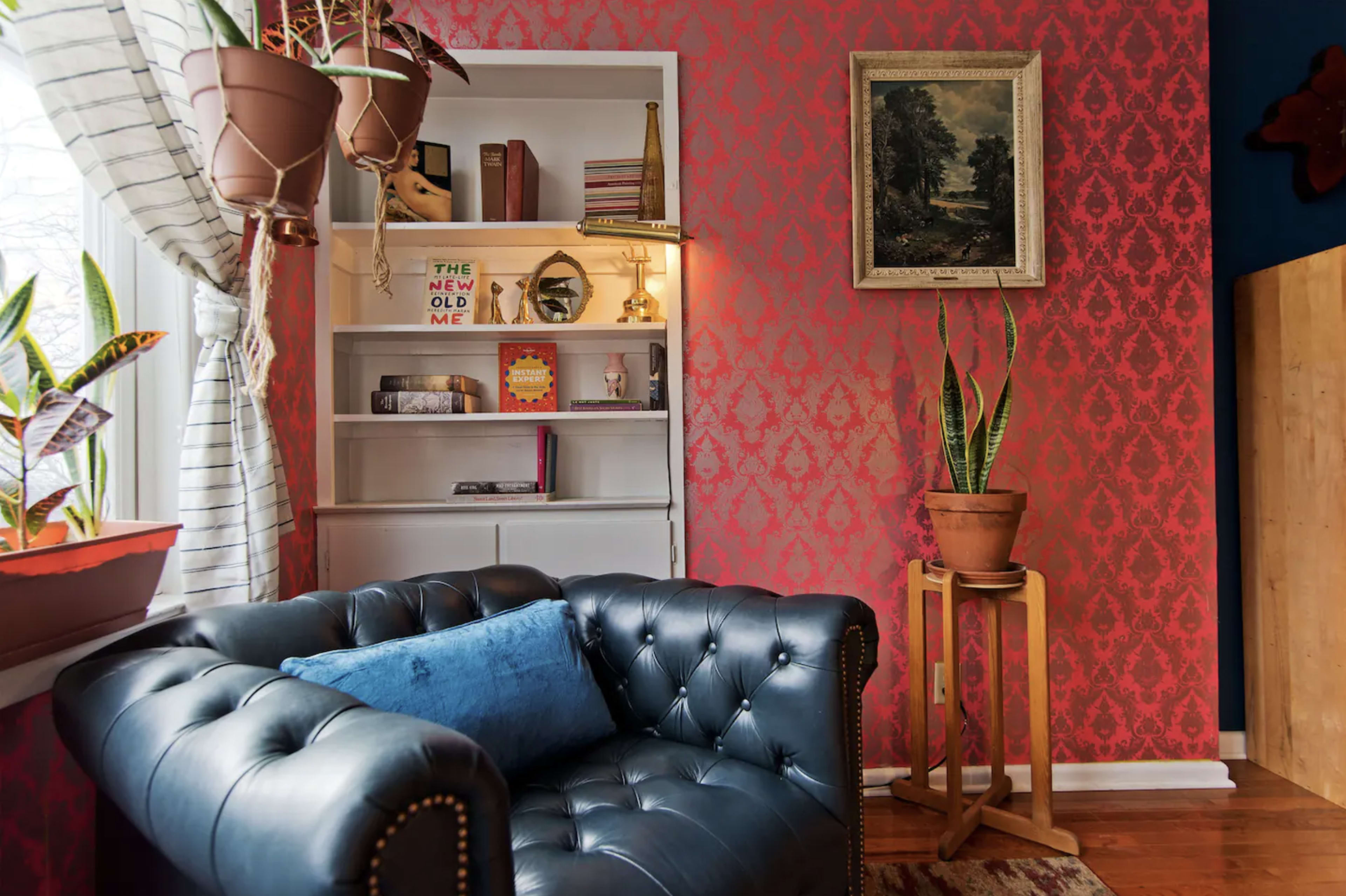 A cozy living space featuring a black leather chesterfield sofa, a patterned red wall, and a white bookshelf adorned with plants and books.
