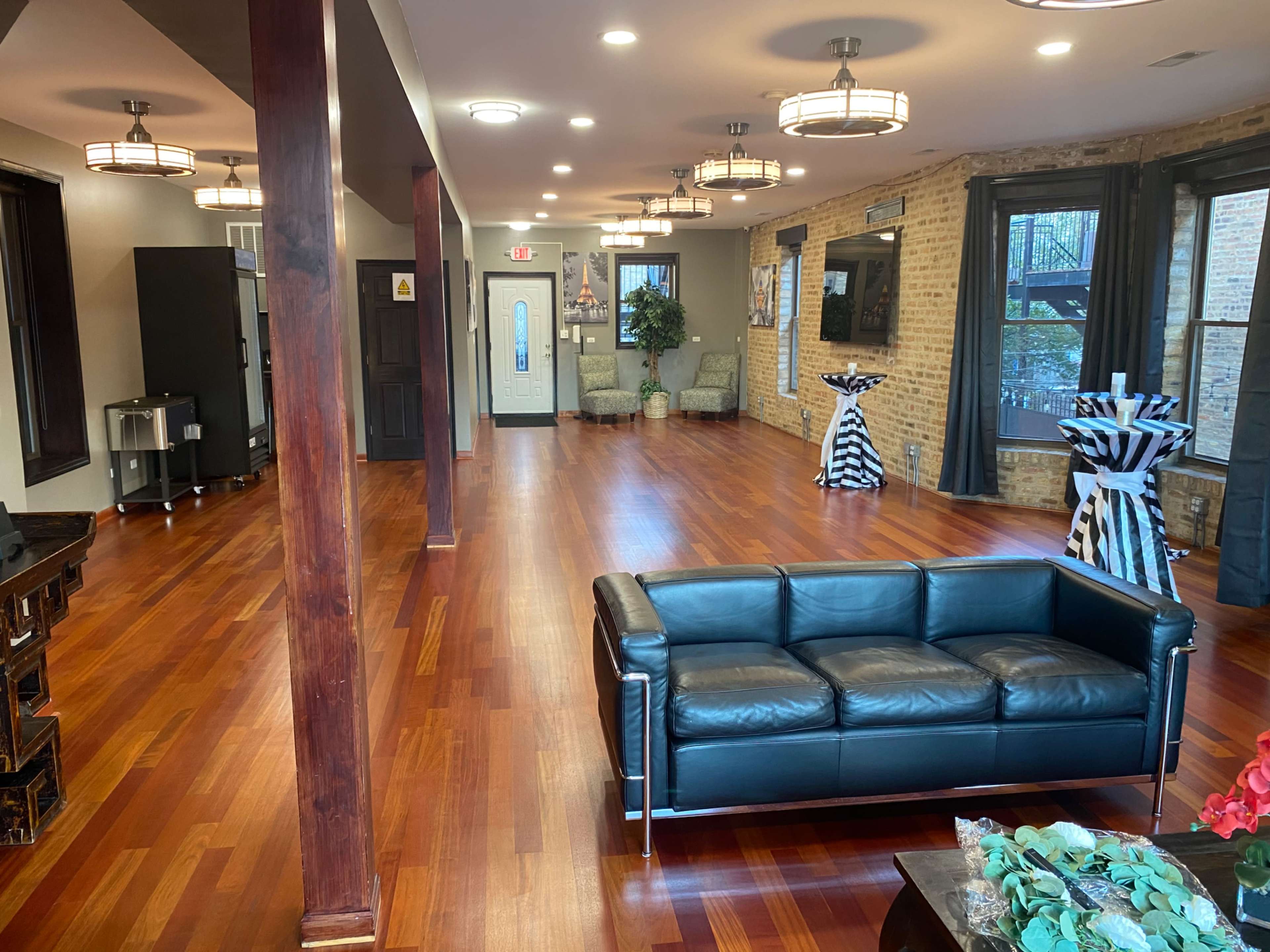 A spacious room with wooden floors, featuring a black leather couch, tables with striped tablecloths, and a seating area near large windows.