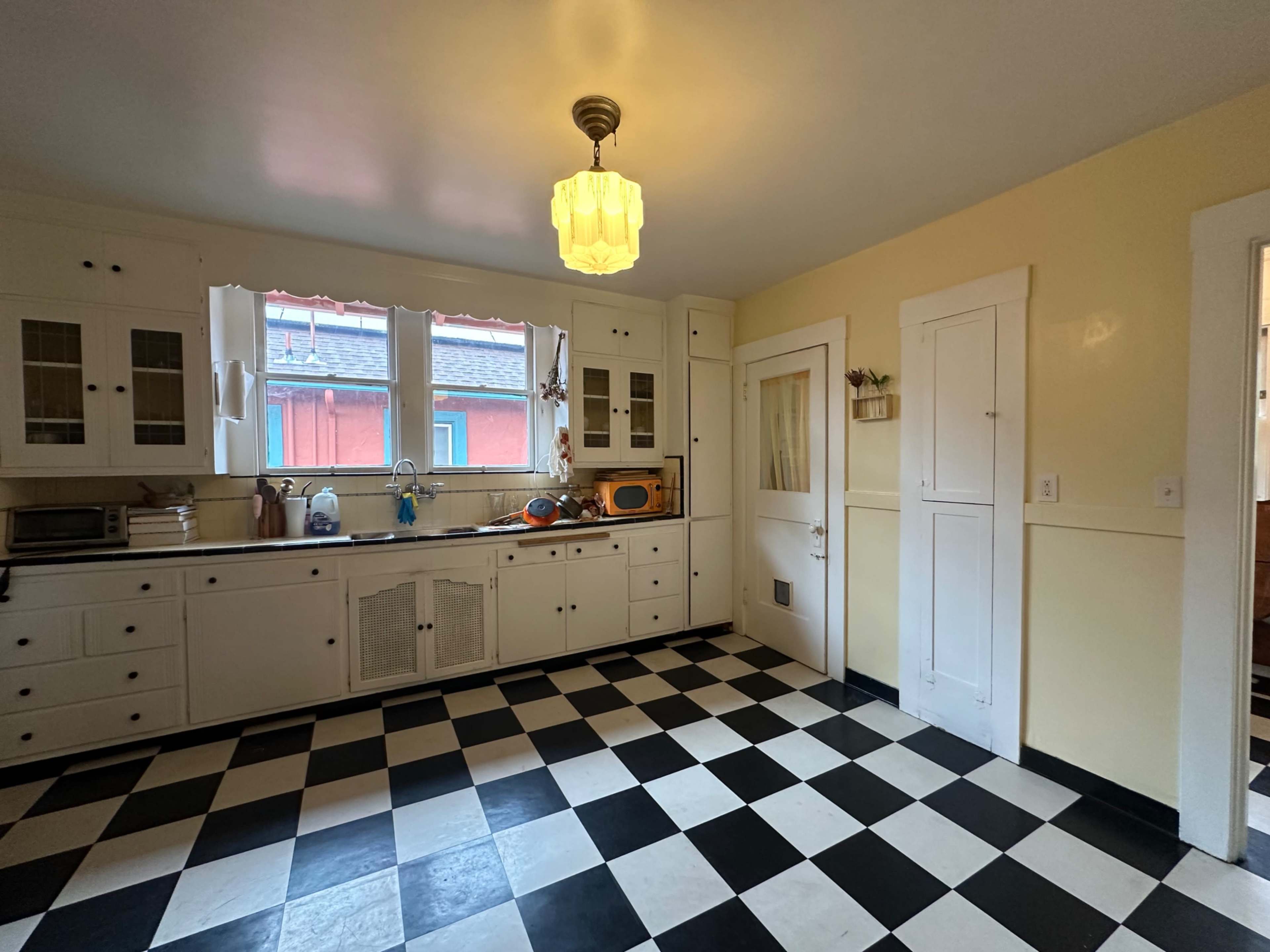 The image shows a kitchen with a black and white checkered floor, white cabinetry, and a vintage light fixture hanging from the ceiling.