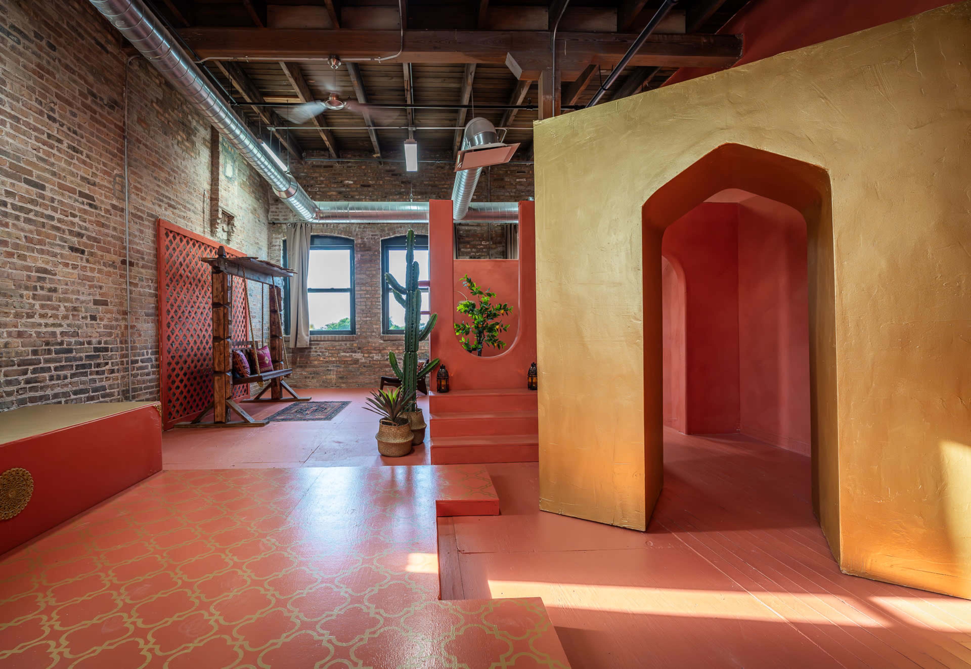 The image shows a vibrant interior space featuring exposed brick walls, colorful painted surfaces, and distinct architectural elements like arches and a cactus in a planter.