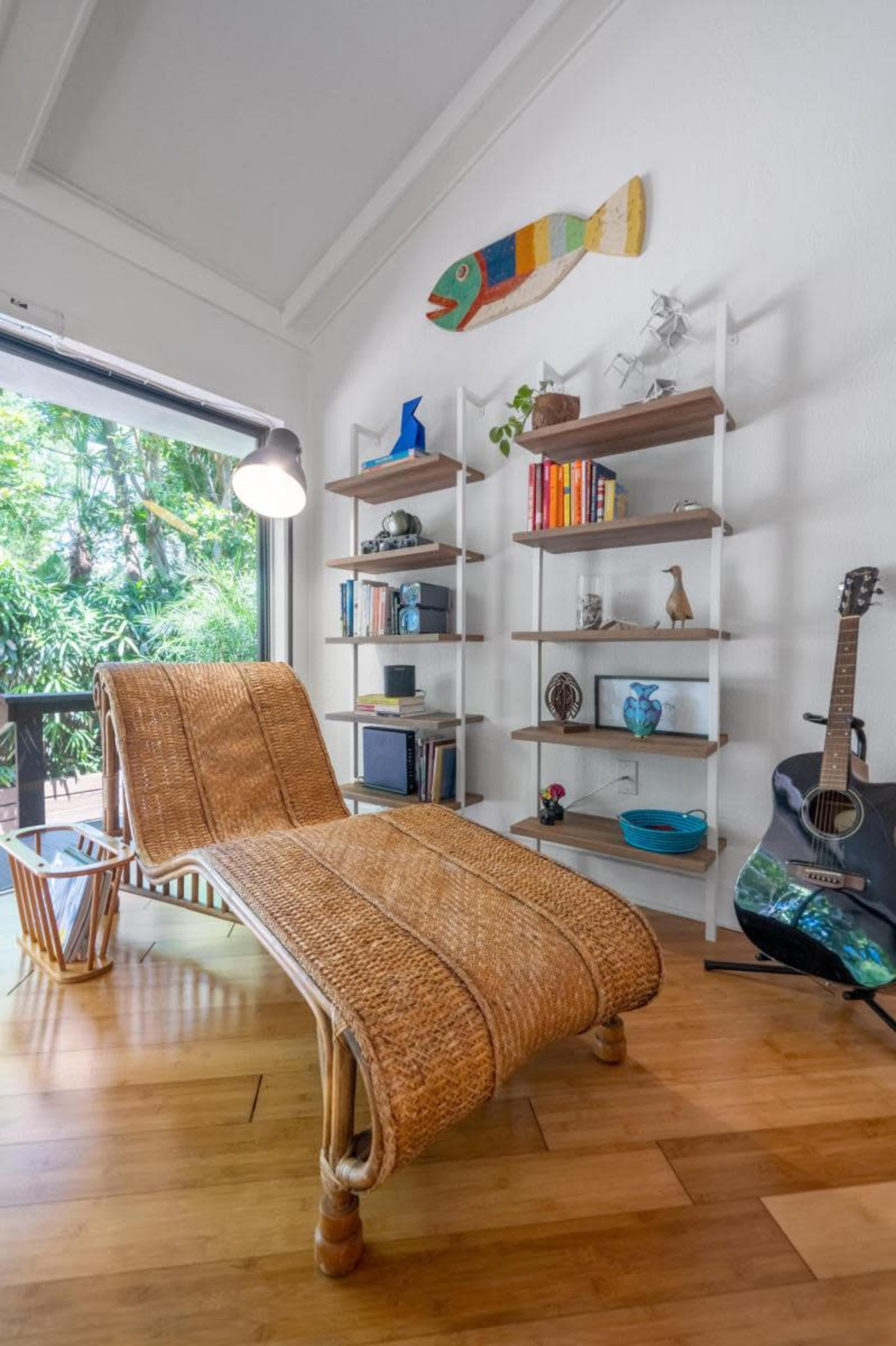 The image shows a cozy indoor space featuring a rattan chaise lounge, a wooden floor, a bookshelf with various decorative items, and a guitar resting against the wall.