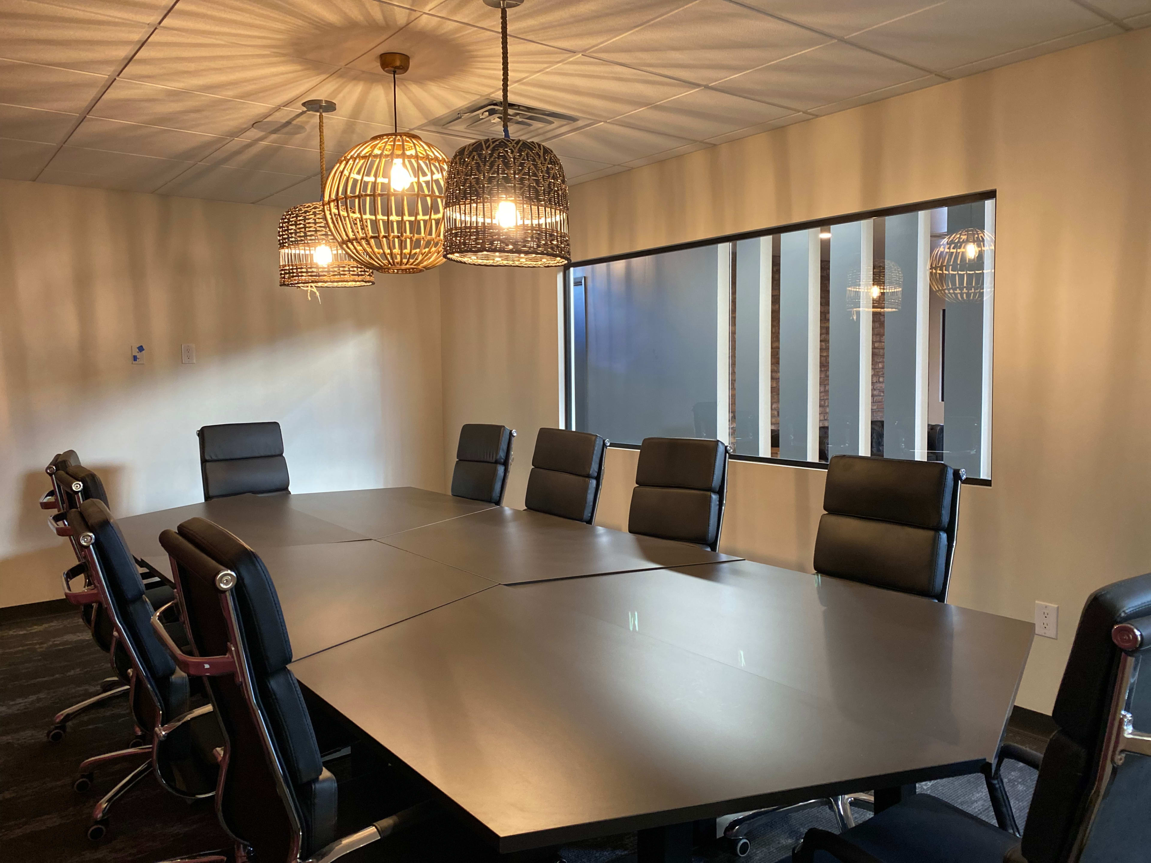 A rectangular conference room features a large table surrounded by eight black leather chairs, with three pendant lights hanging from the ceiling and a window providing a view of a hallway.