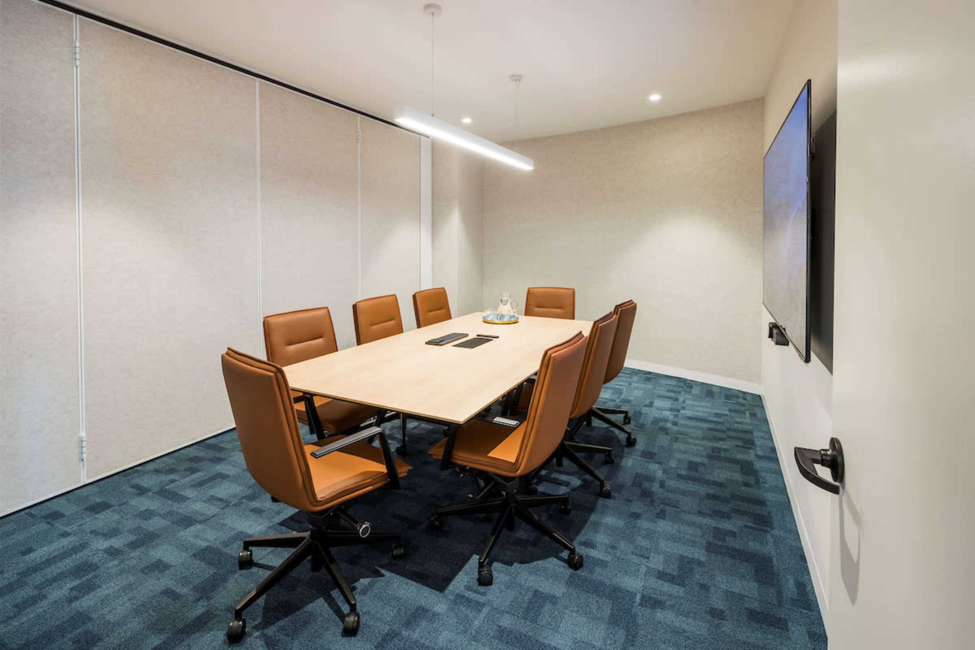 A well-lit meeting room features a large wooden table surrounded by eight brown leather chairs, with a screen mounted on one wall.