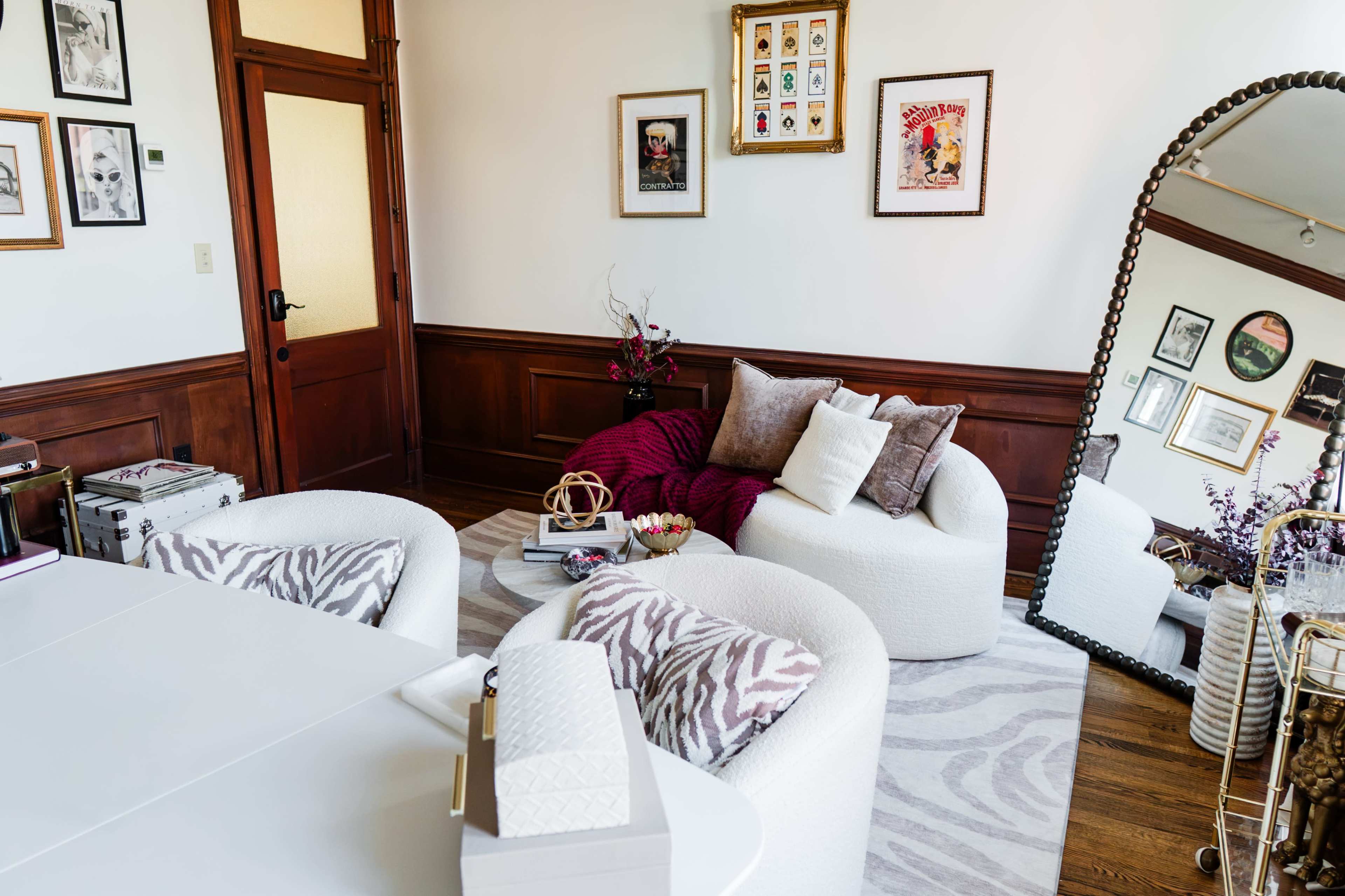 The image shows a cozy sitting area featuring two white upholstered chairs, a round coffee table, and a large mirror on the wall, all set against a backdrop of framed artwork and wooden paneling.