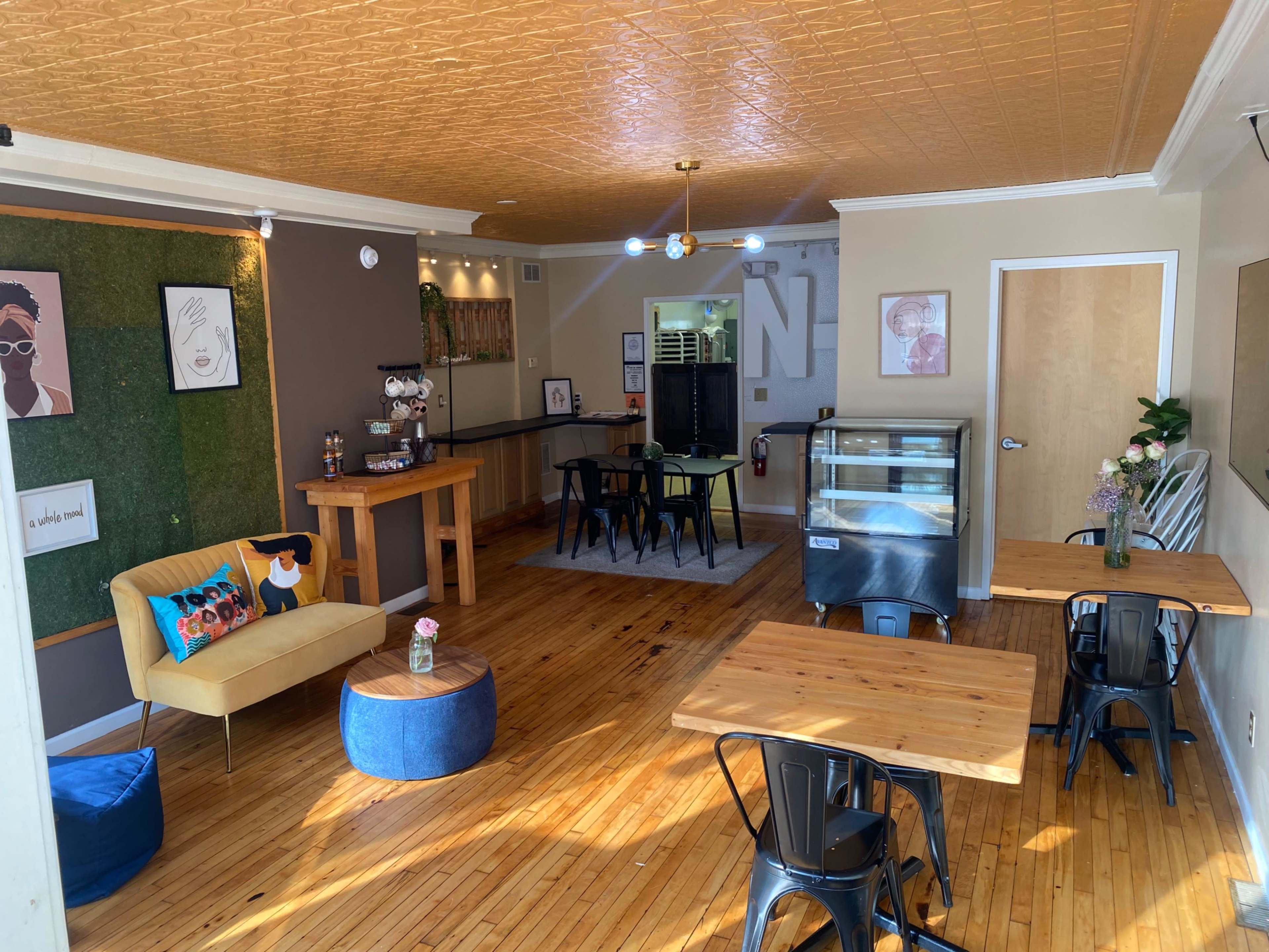 The image shows a cozy café interior with wooden floors, a mix of seating options, and a display case in the background.