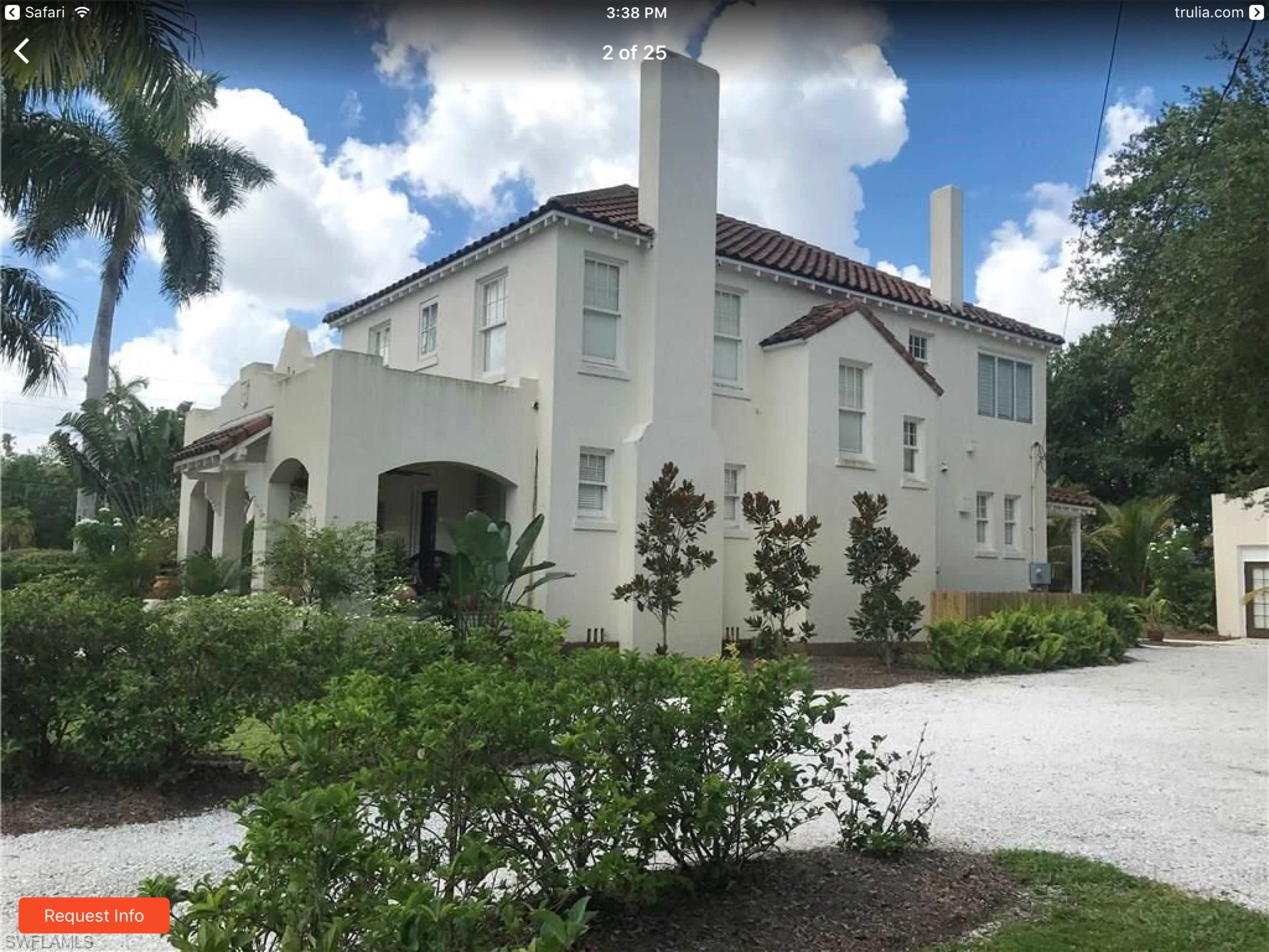 A two-story, white stucco house with a tile roof and surrounding greenery is situated on a gravel driveway.