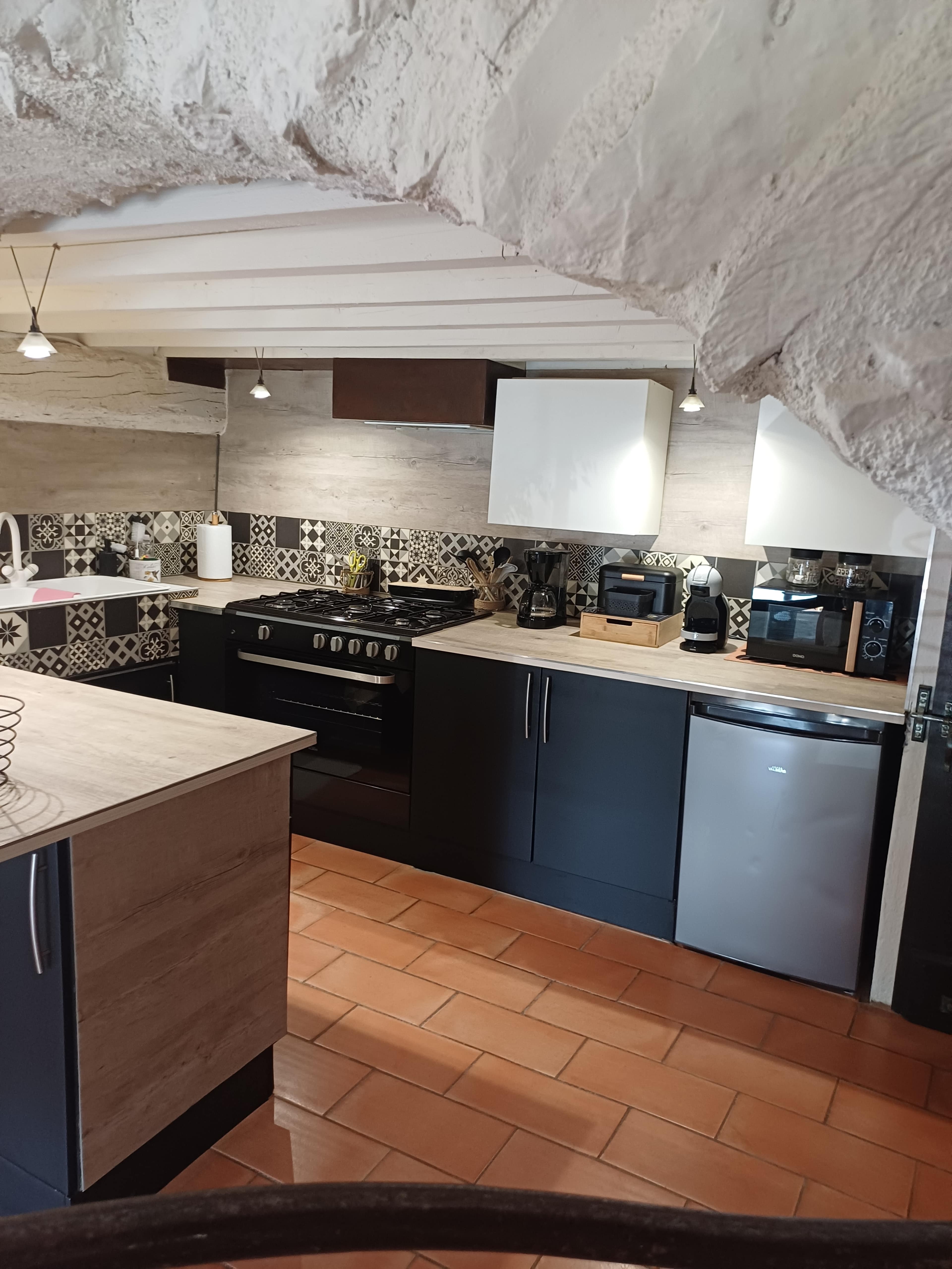 The image shows a modern kitchen featuring a mix of dark cabinetry, a stone wall, and patterned tile backsplash, with various appliances and a large oven.