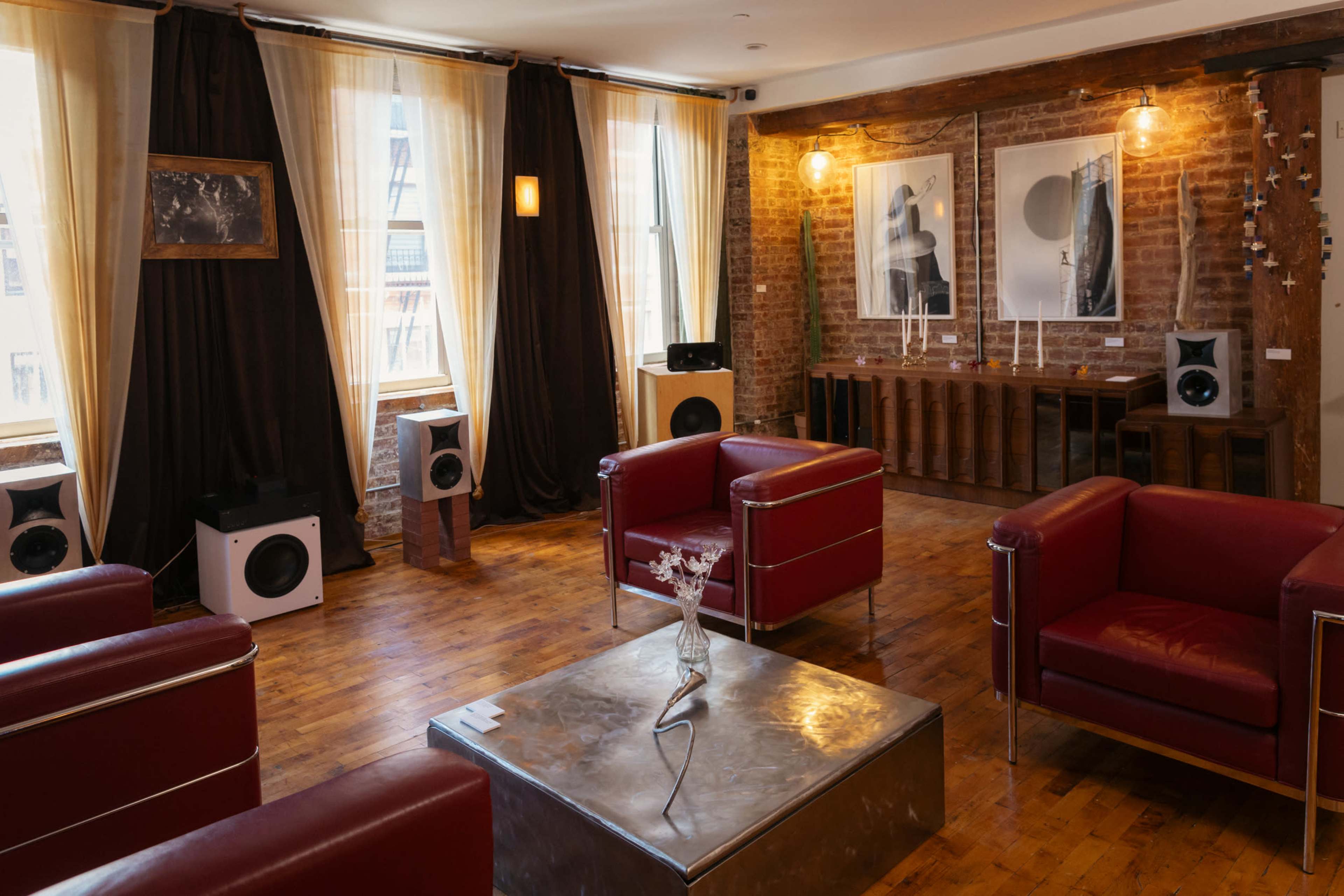 The image shows a modern interior space with red leather chairs, a metal table, and speakers positioned around a room with exposed brick walls and large windows.