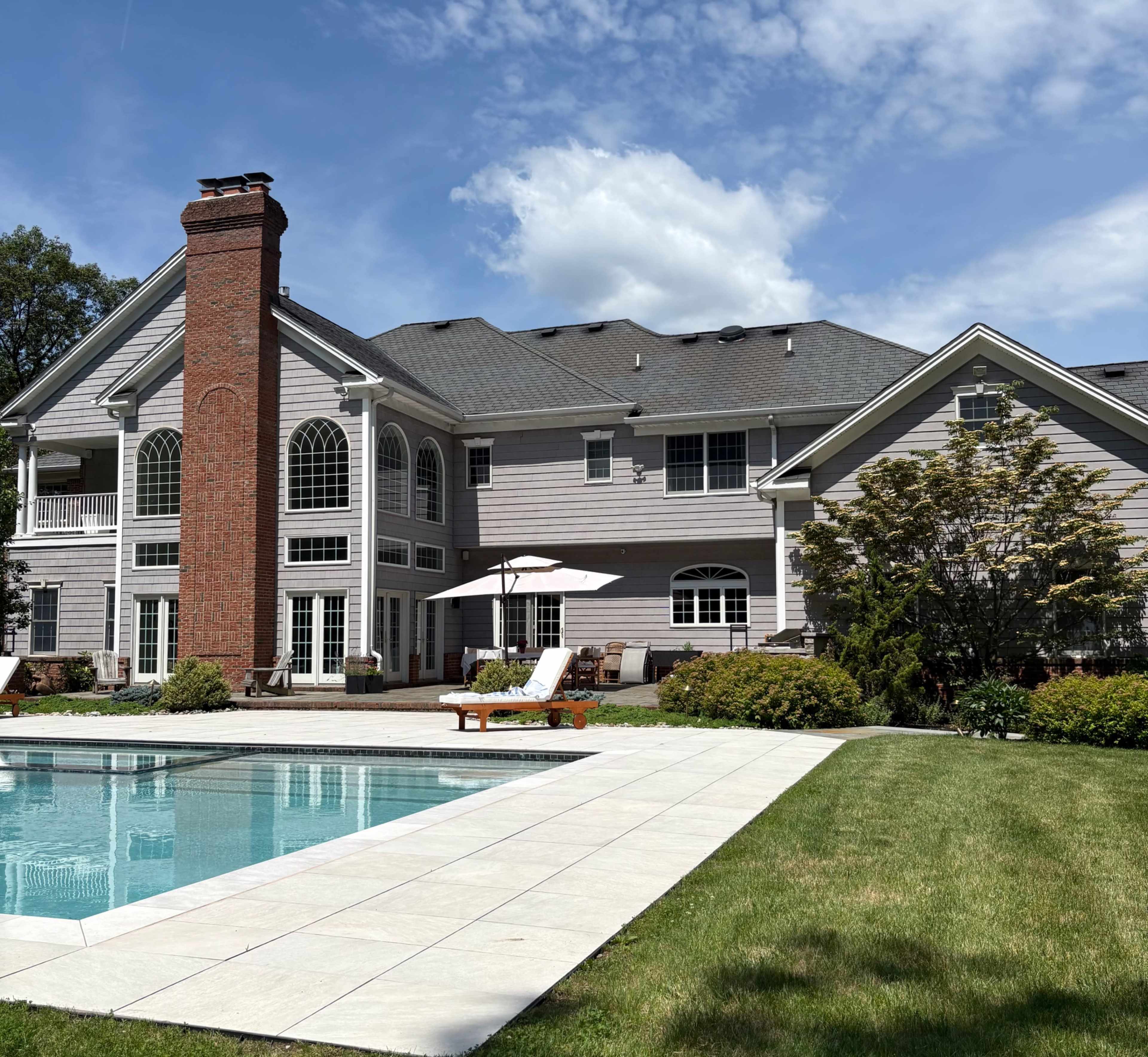 The image features a two-story house with a brick chimney, surrounded by a pool and landscaped yard under a partly cloudy sky.