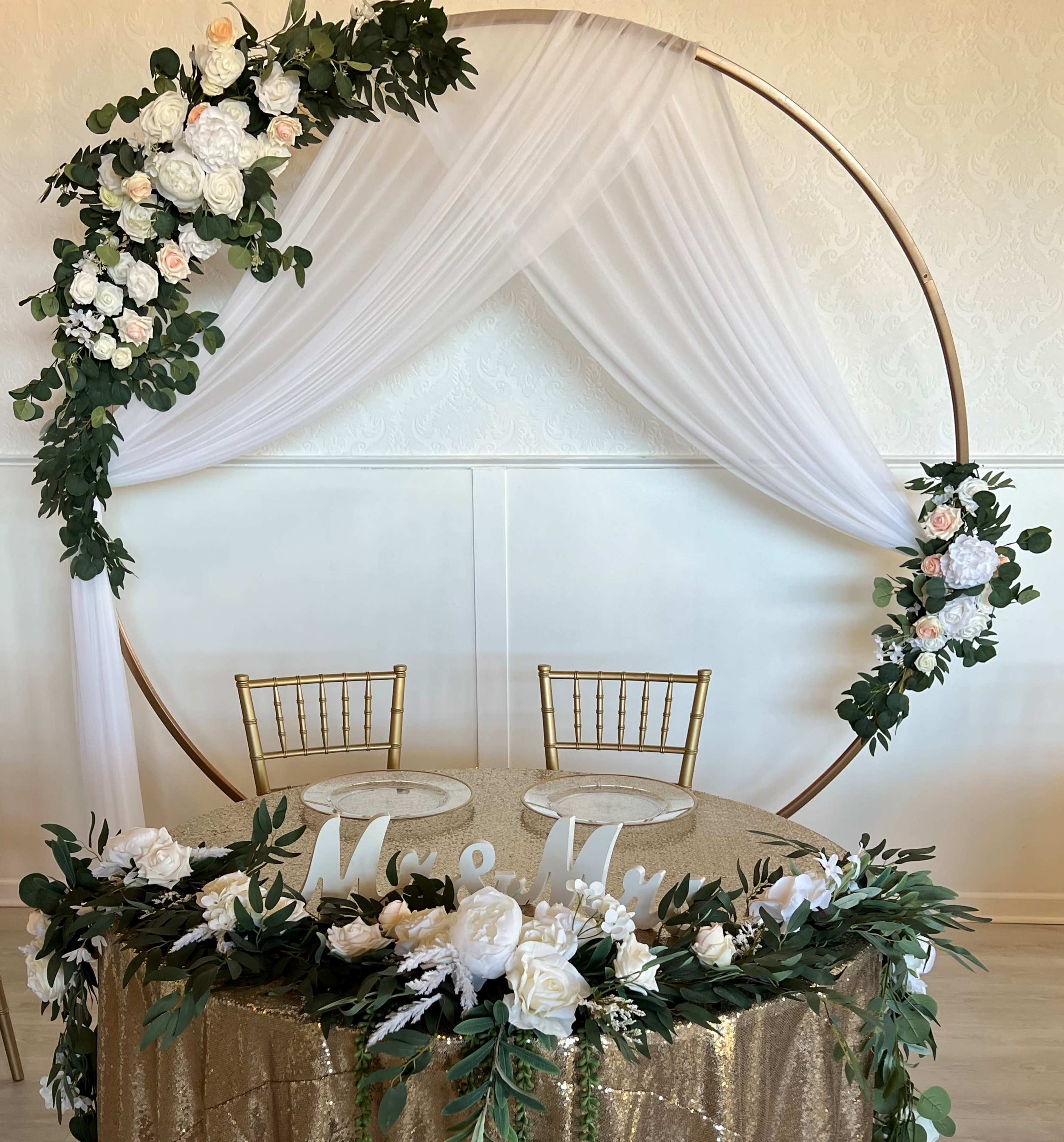 A round floral arch is decorated with white and pink flowers, framing a table with a gold sequin cloth and two empty gold chairs.