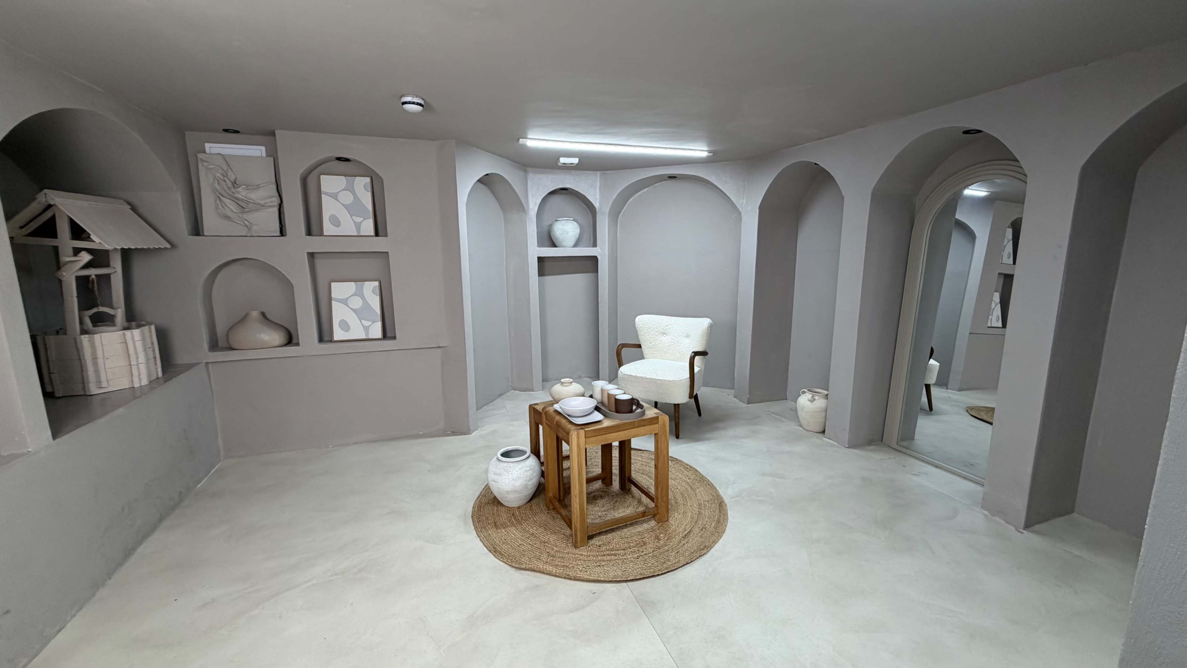 The image shows a modern, minimalist room featuring arches, decorative shelves, a white chair, a wooden table, and various ceramics arranged aesthetically.