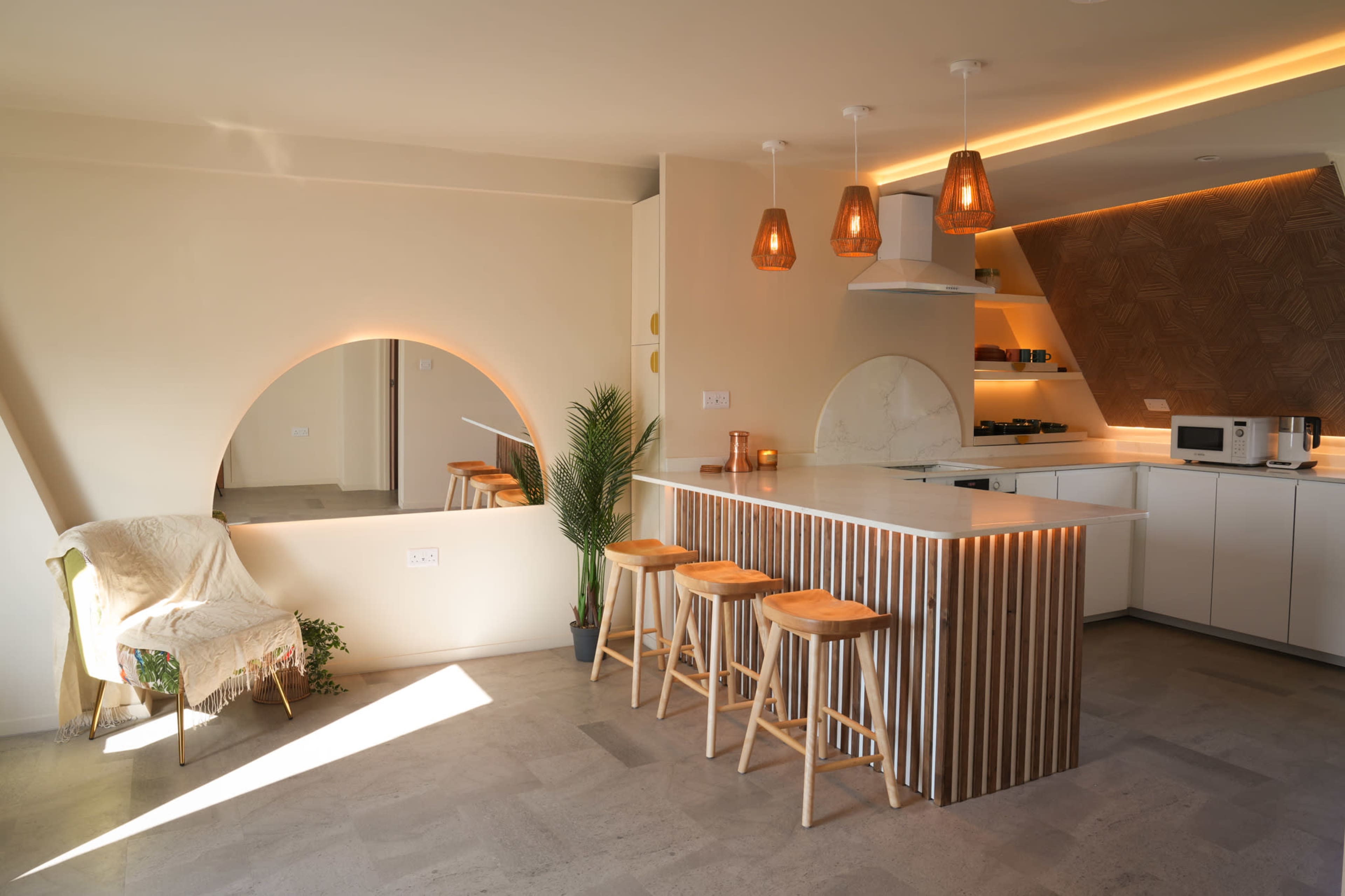 A modern kitchen with a central island made of slatted wood, three wooden stools, and pendant lighting, complemented by a curved archway and a decorative plant.