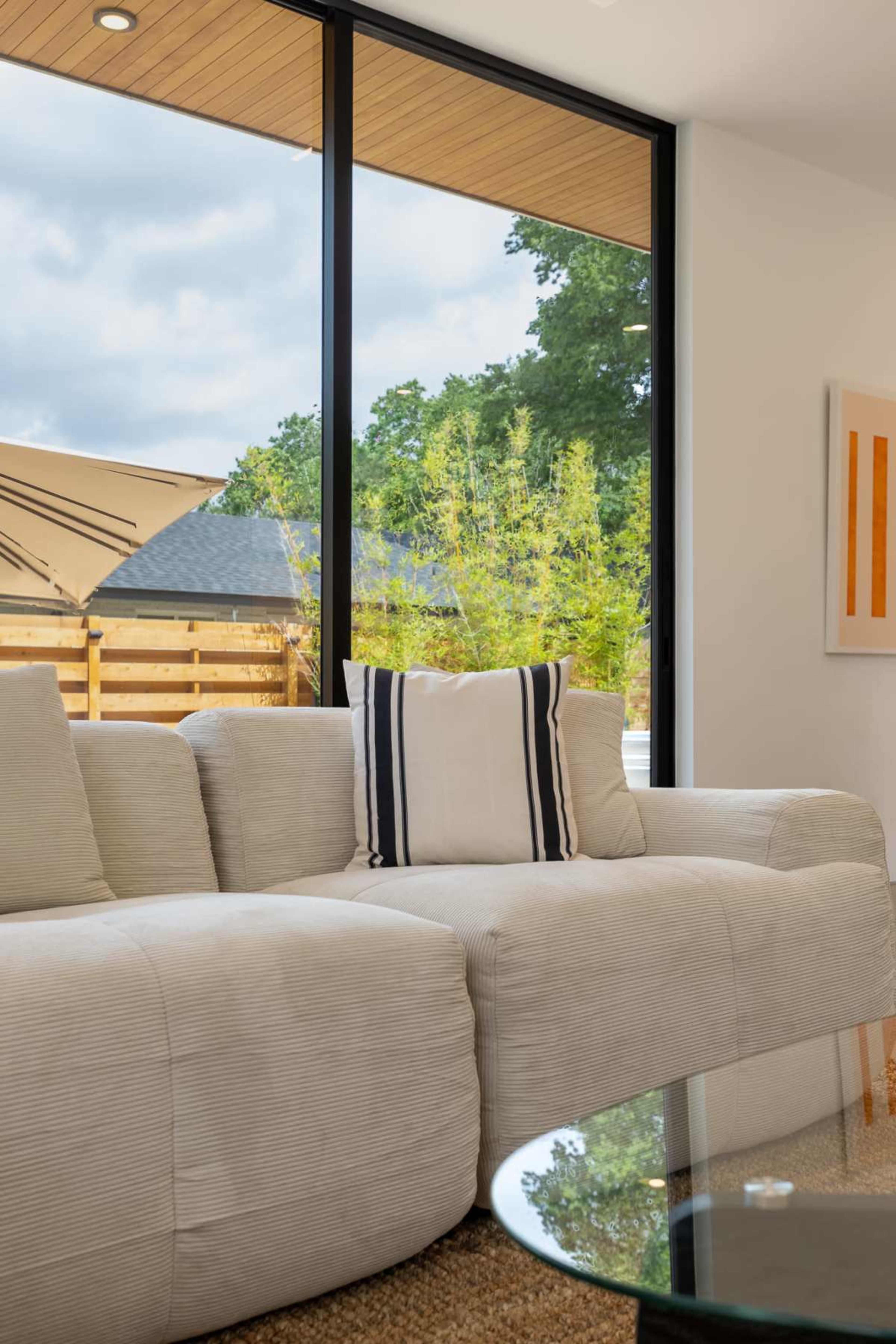 The image shows a modern living room with a light-colored sofa, a glass coffee table, and large windows revealing greenery outside.