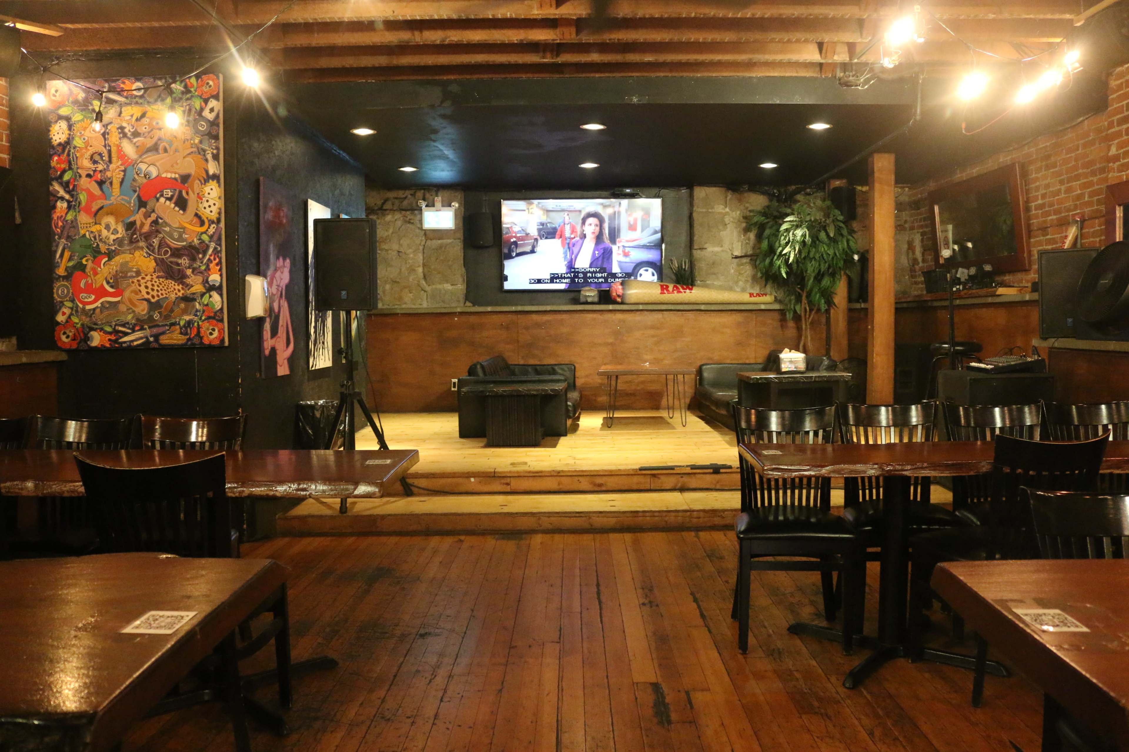 A dimly lit bar or venue with wooden floors, a stage in the center, tables arranged in front, and a television mounted on the wall.
