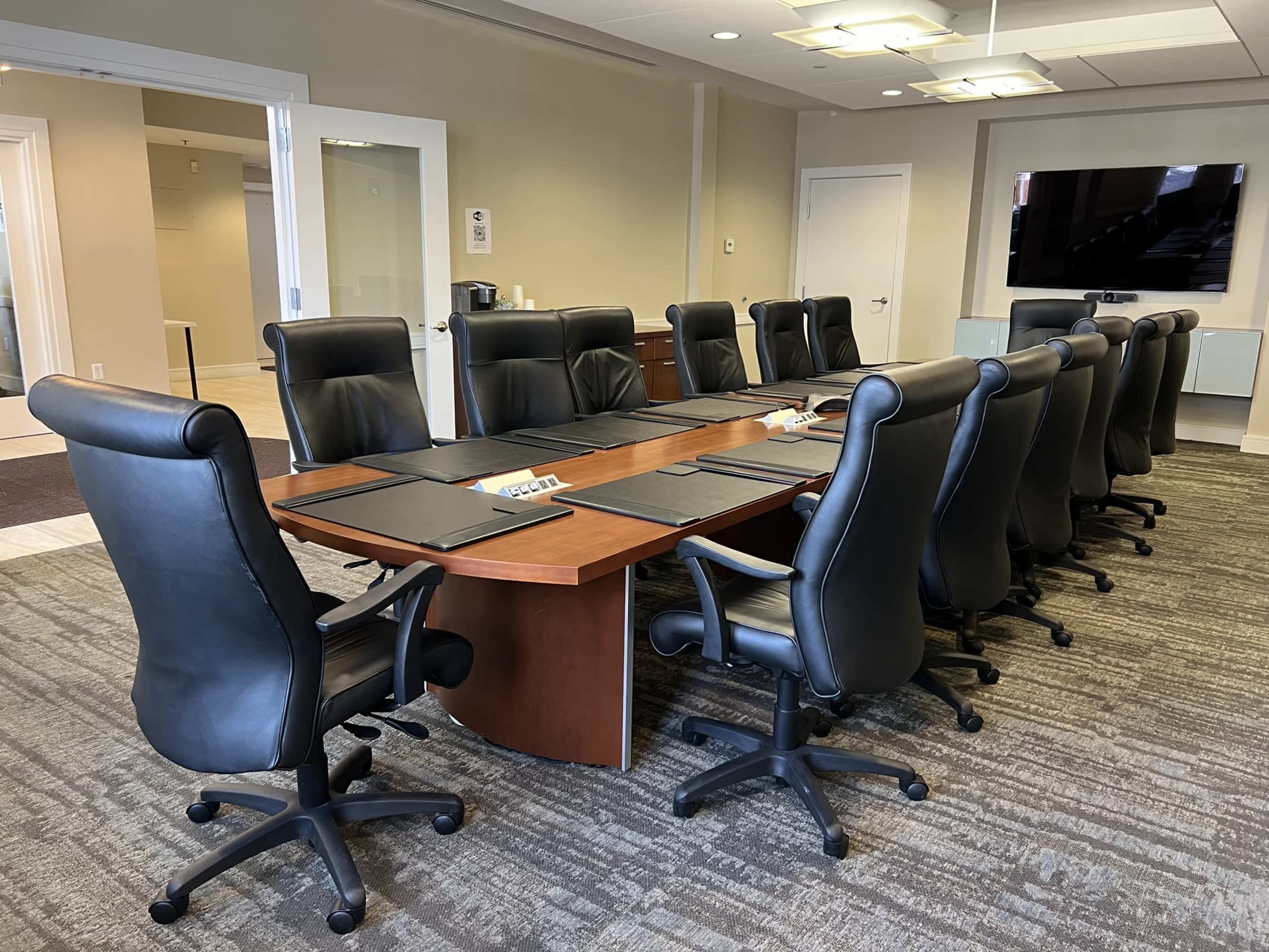 A conference room featuring a long wooden table surrounded by ergonomic black office chairs and a large television on the wall.