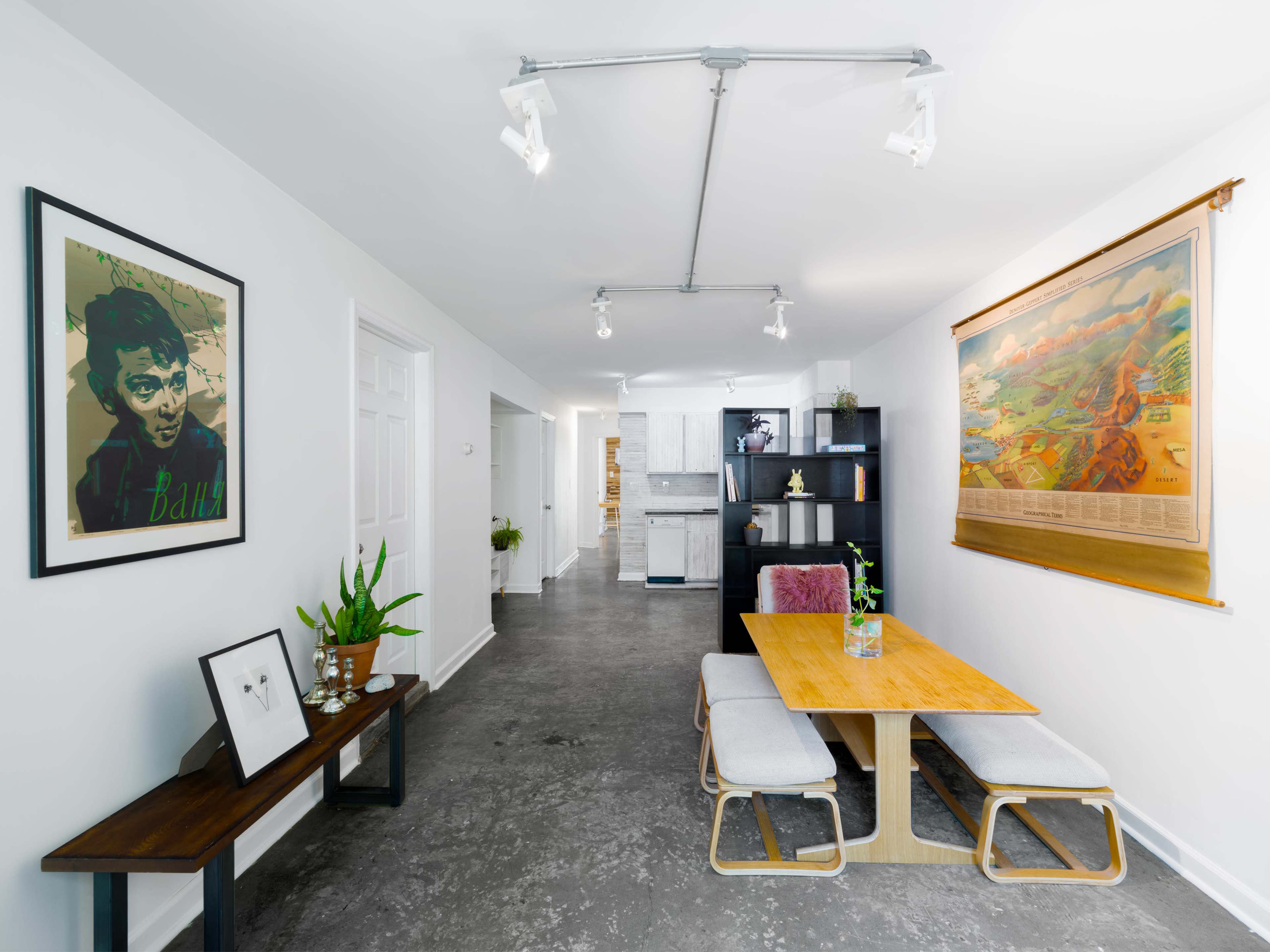 The image shows a modern dining area with a wooden table, gray upholstered chairs, artwork on the walls, and a bookshelf against one side.