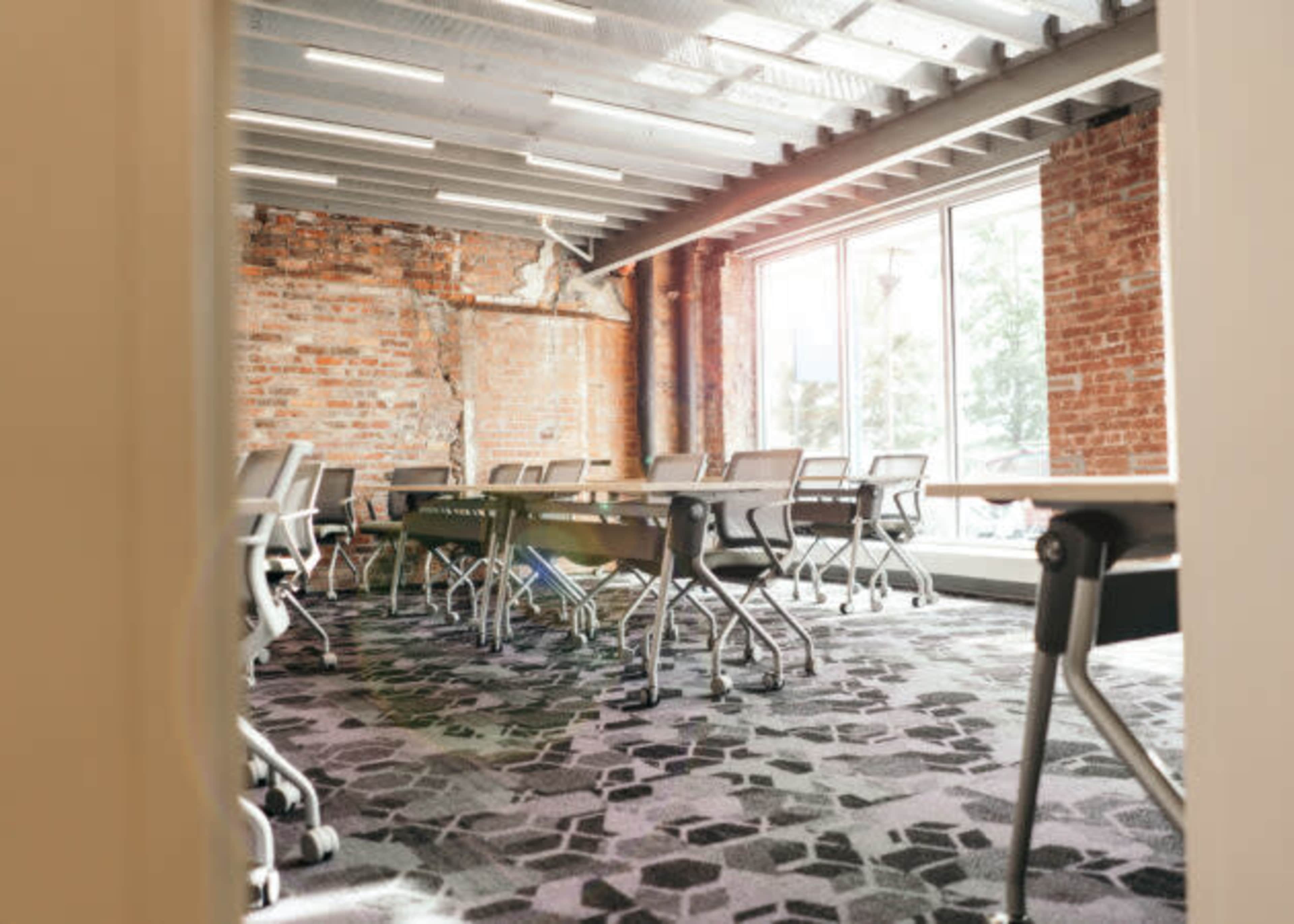 The image shows a modern conference room with a large table and several chairs arranged around it, featuring exposed brick walls and large windows.