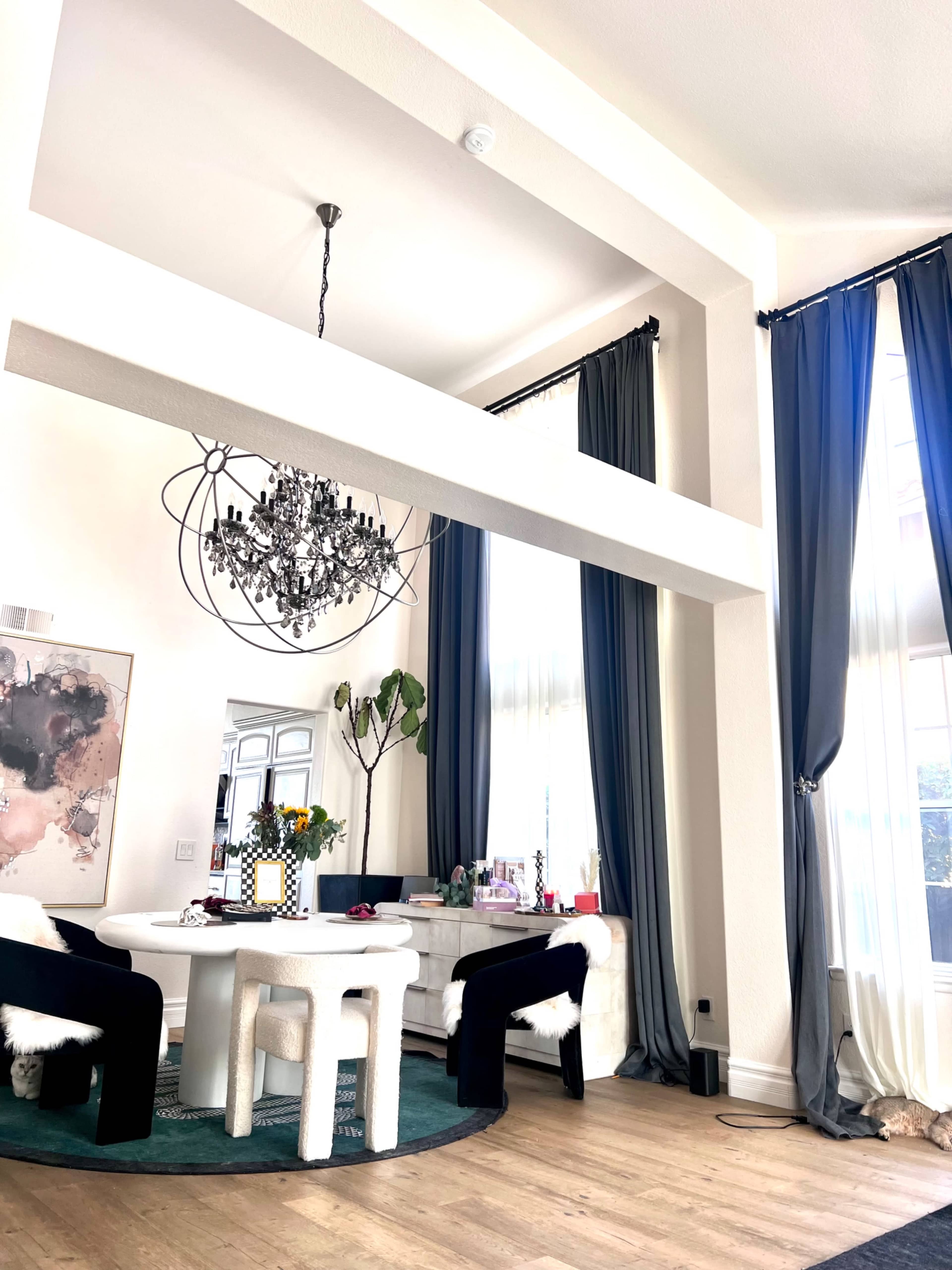 A modern dining area features a round table with black chairs, a decorative chandelier overhead, and large windows draped with blue and gray curtains.