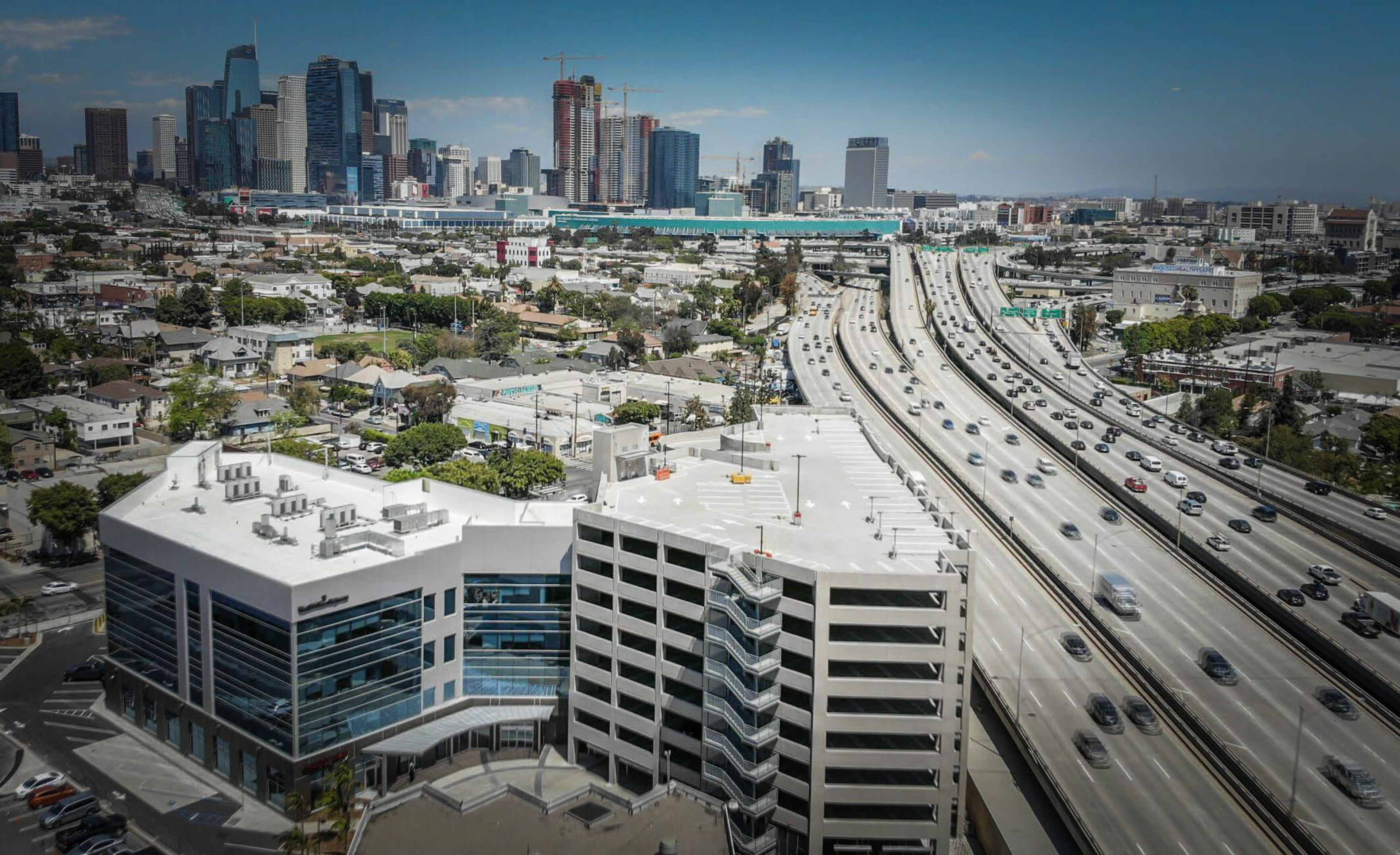 The image shows a view of a city skyline with tall buildings in the background, an extensive highway with moving traffic in the foreground, and a modern building adjacent to the road.