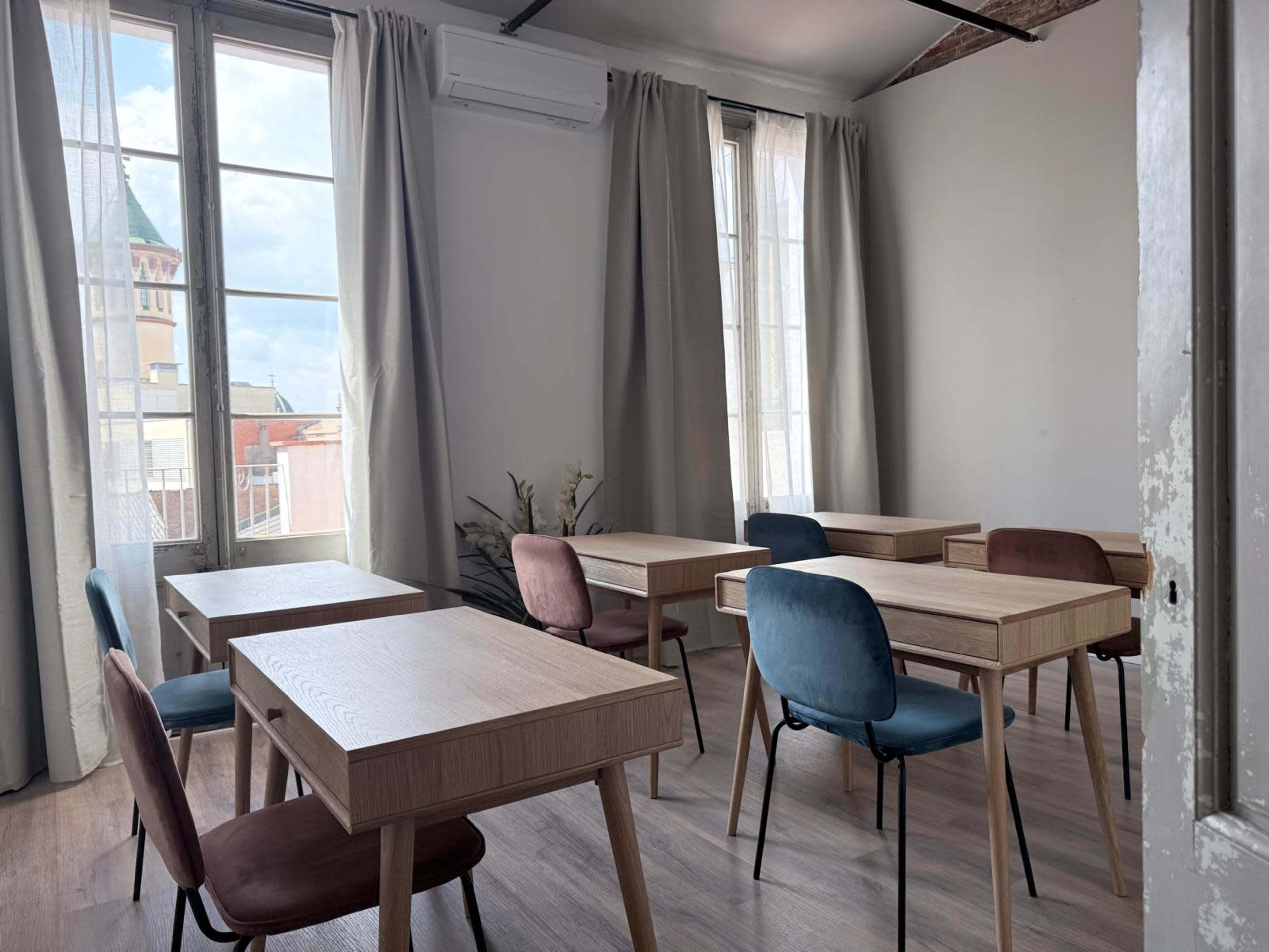 A bright, minimalist room with several wooden tables and chairs arranged near large windows, allowing natural light to enter.