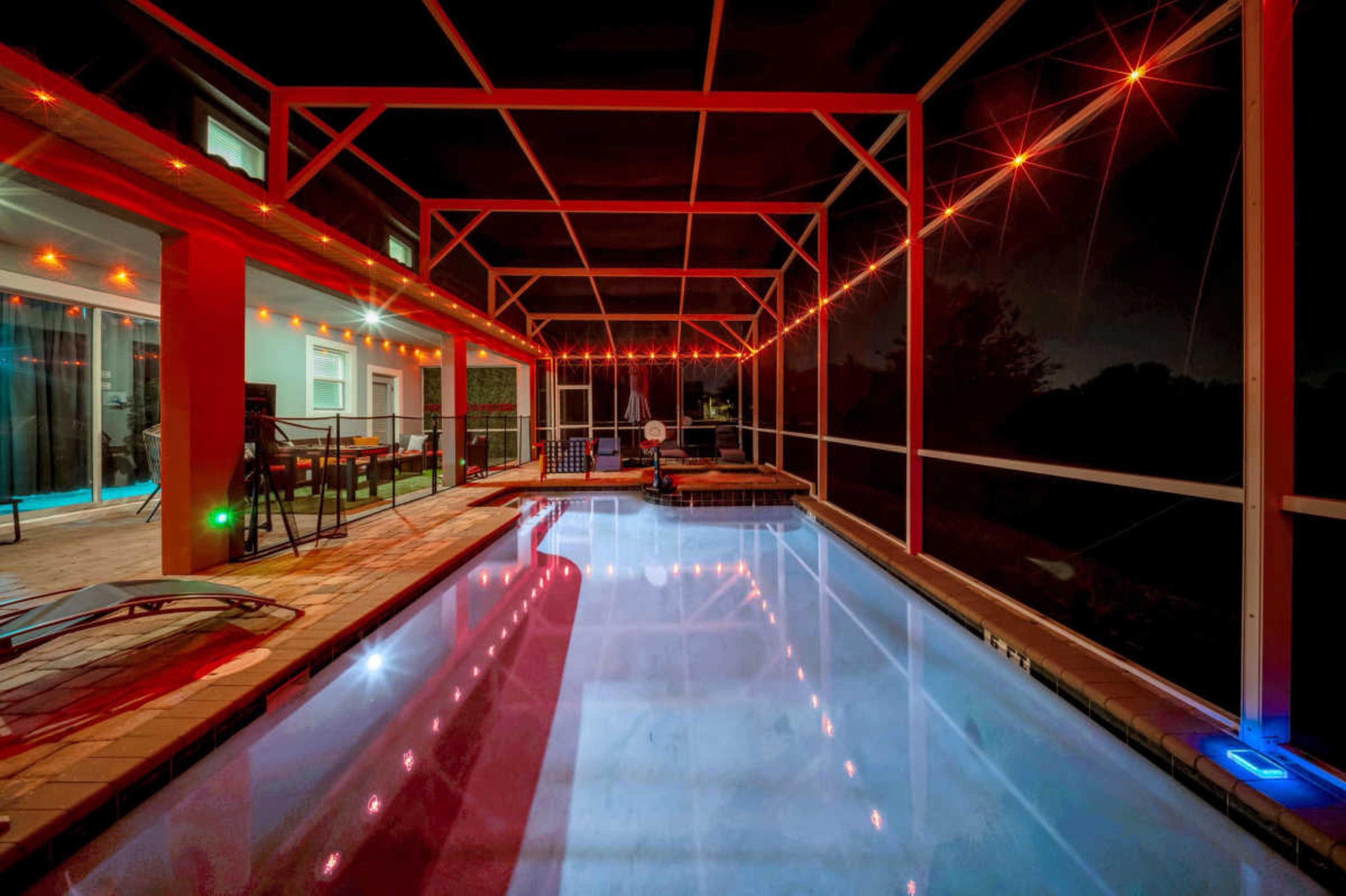 The image shows a screened-in pool area at night, illuminated by string lights and featuring a patio with chairs and a table.
