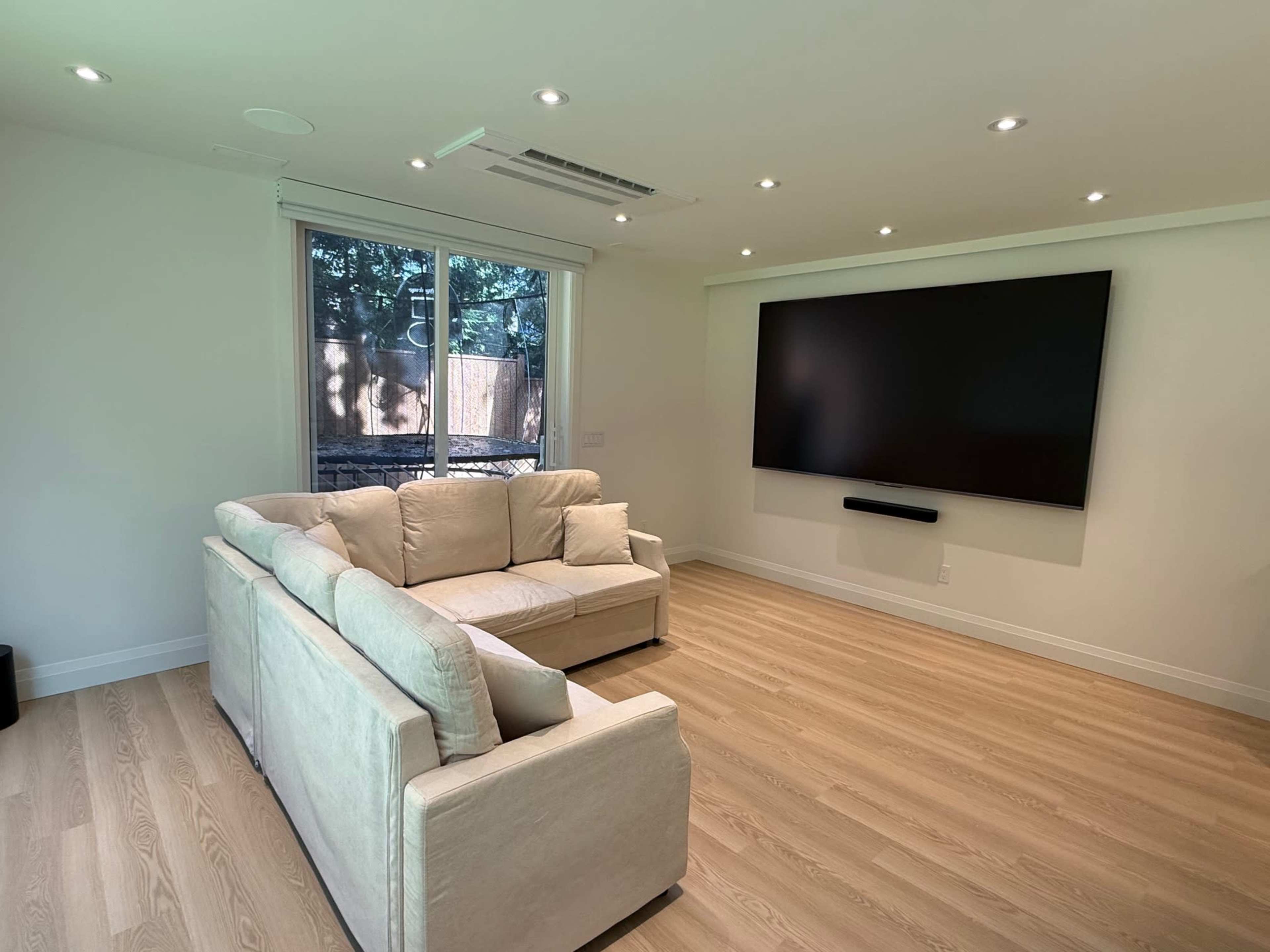 The image shows a modern living room with a white sofa facing a mounted flat-screen TV on a light-colored wall, illuminated by recessed lighting.
