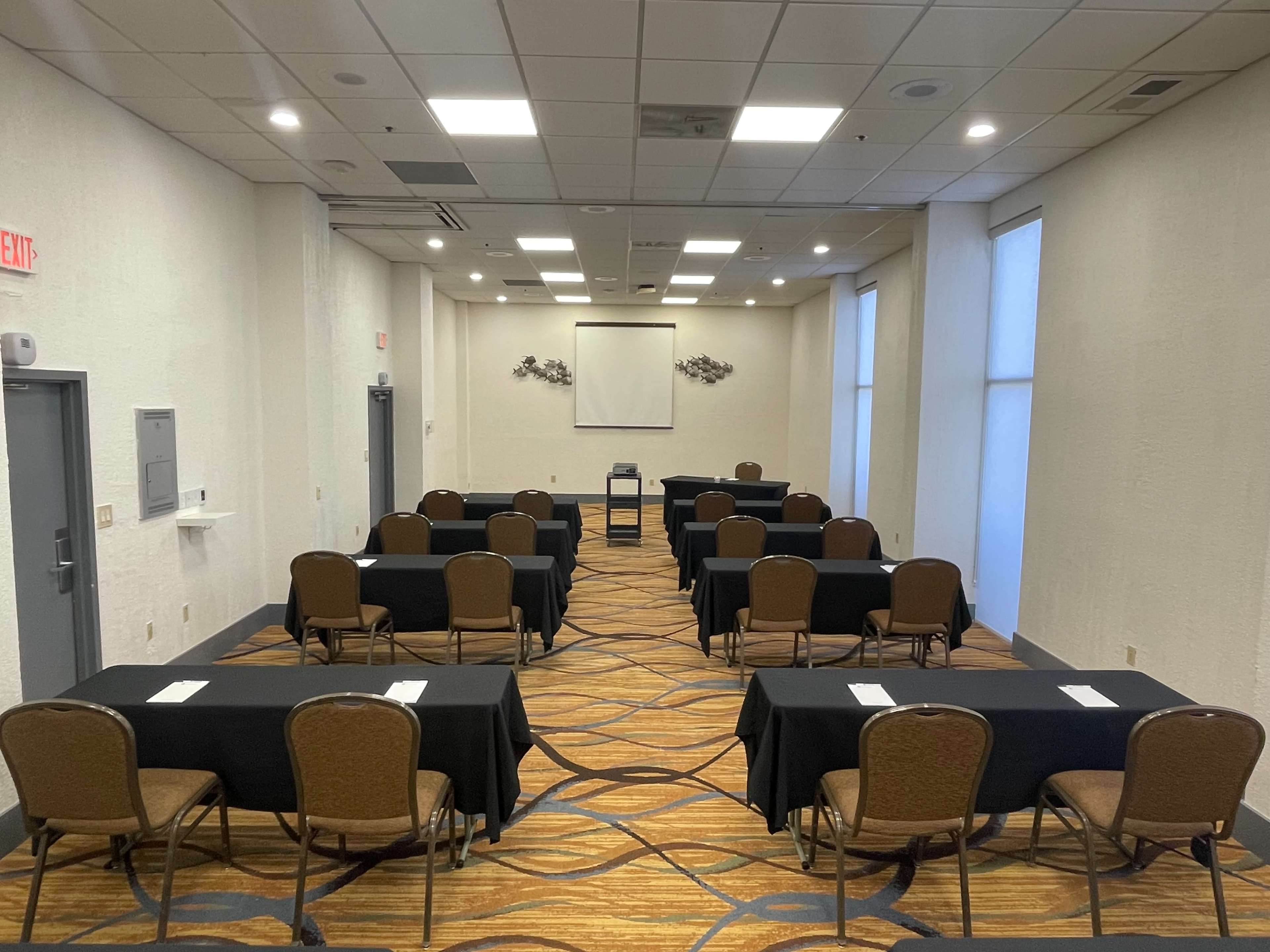 A conference room setup with rows of tables and chairs arranged for a meeting, featuring a projector screen at the front.