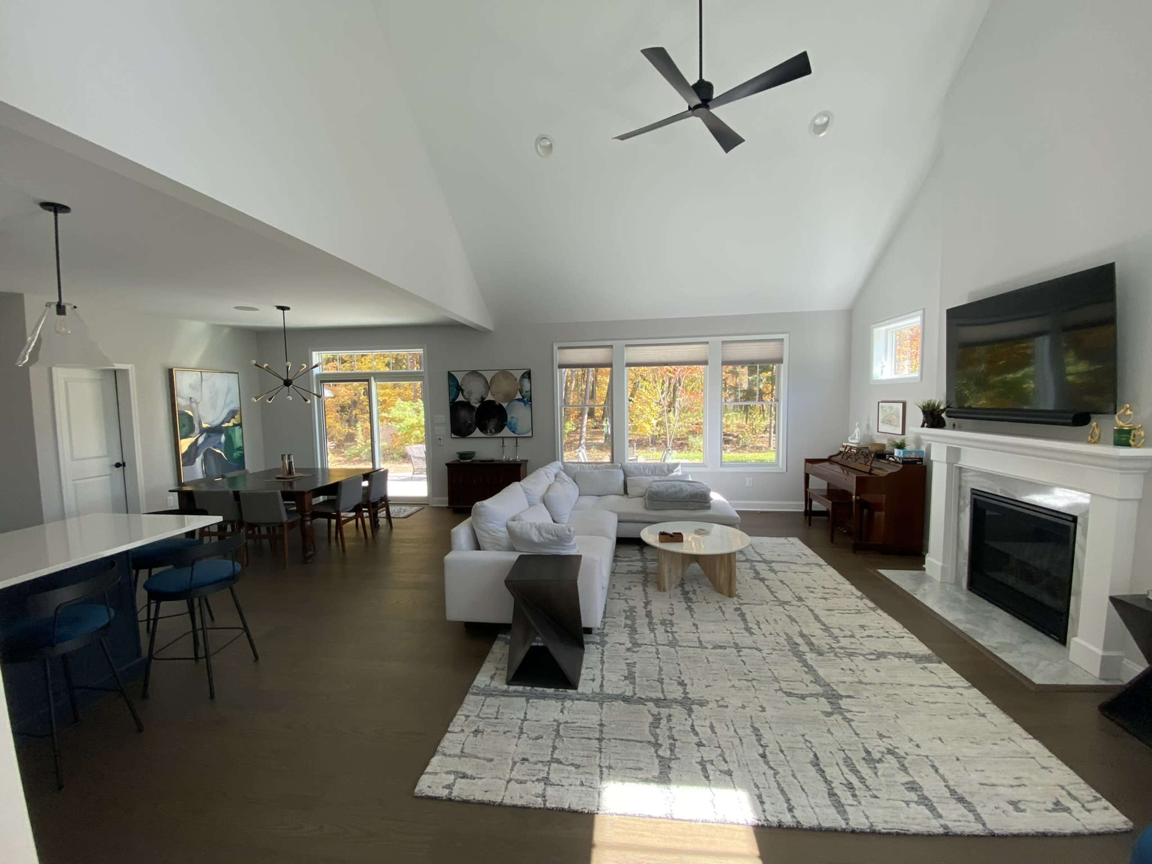 A modern open-concept living room features a white sectional sofa, a wooden coffee table, and large windows overlooking a tree-filled view.