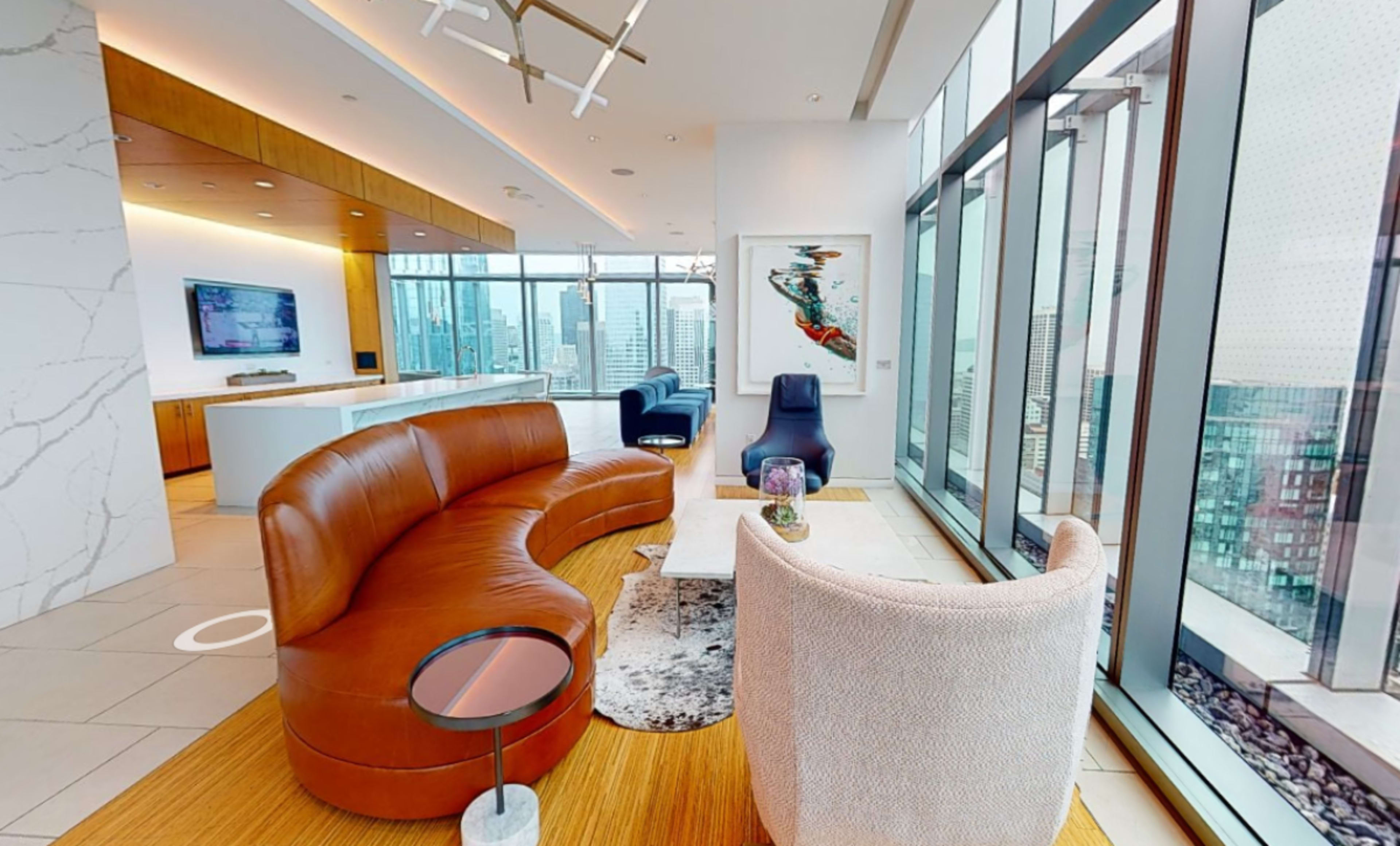 A modern, open-concept living space with large windows offering city views, featuring a curved brown leather sofa, a white armchair, and a central coffee table on a wooden floor.