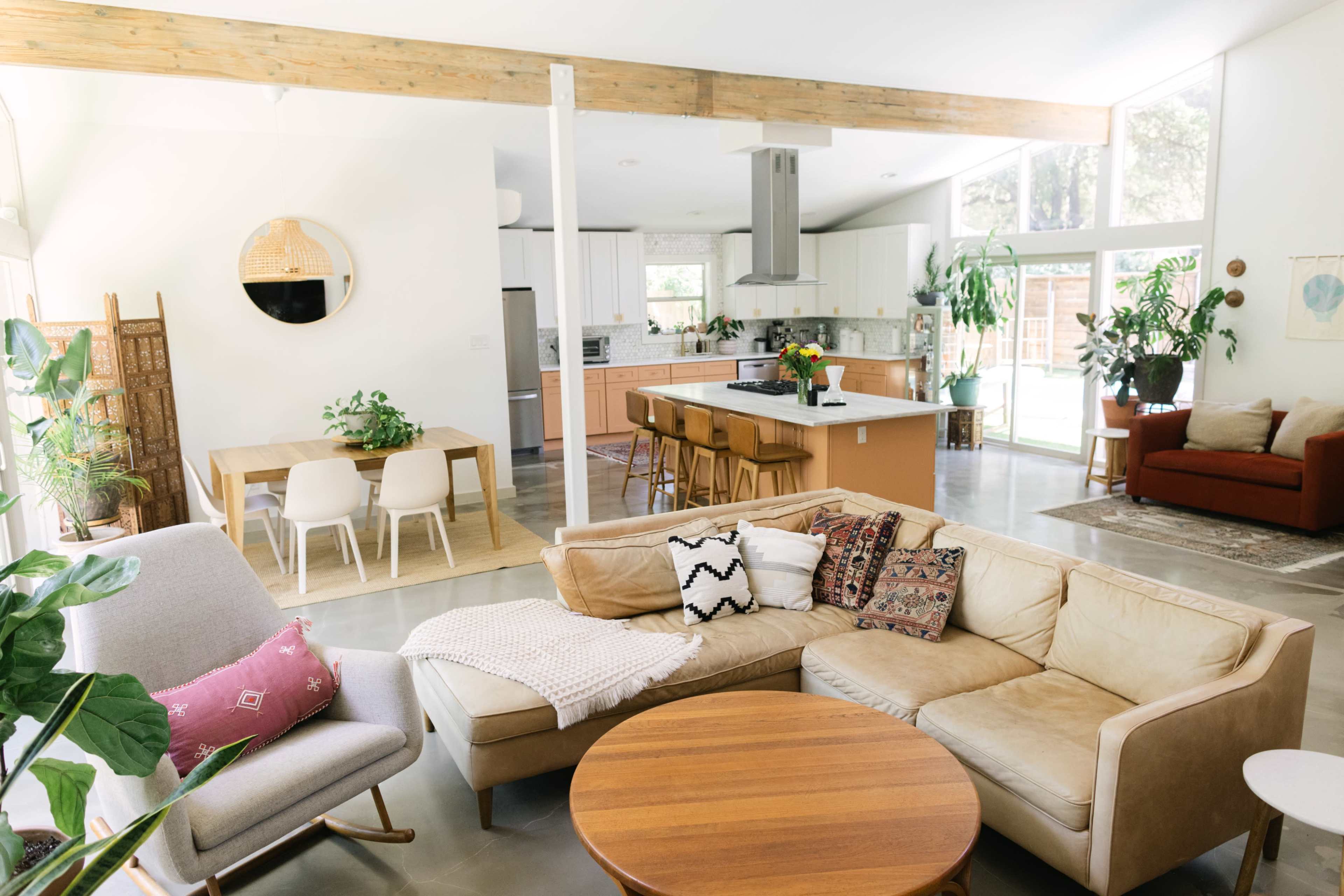 A modern, open-concept living space featuring a beige sectional sofa, a round wooden coffee table, a dining area with white chairs, and a kitchen with a central island.