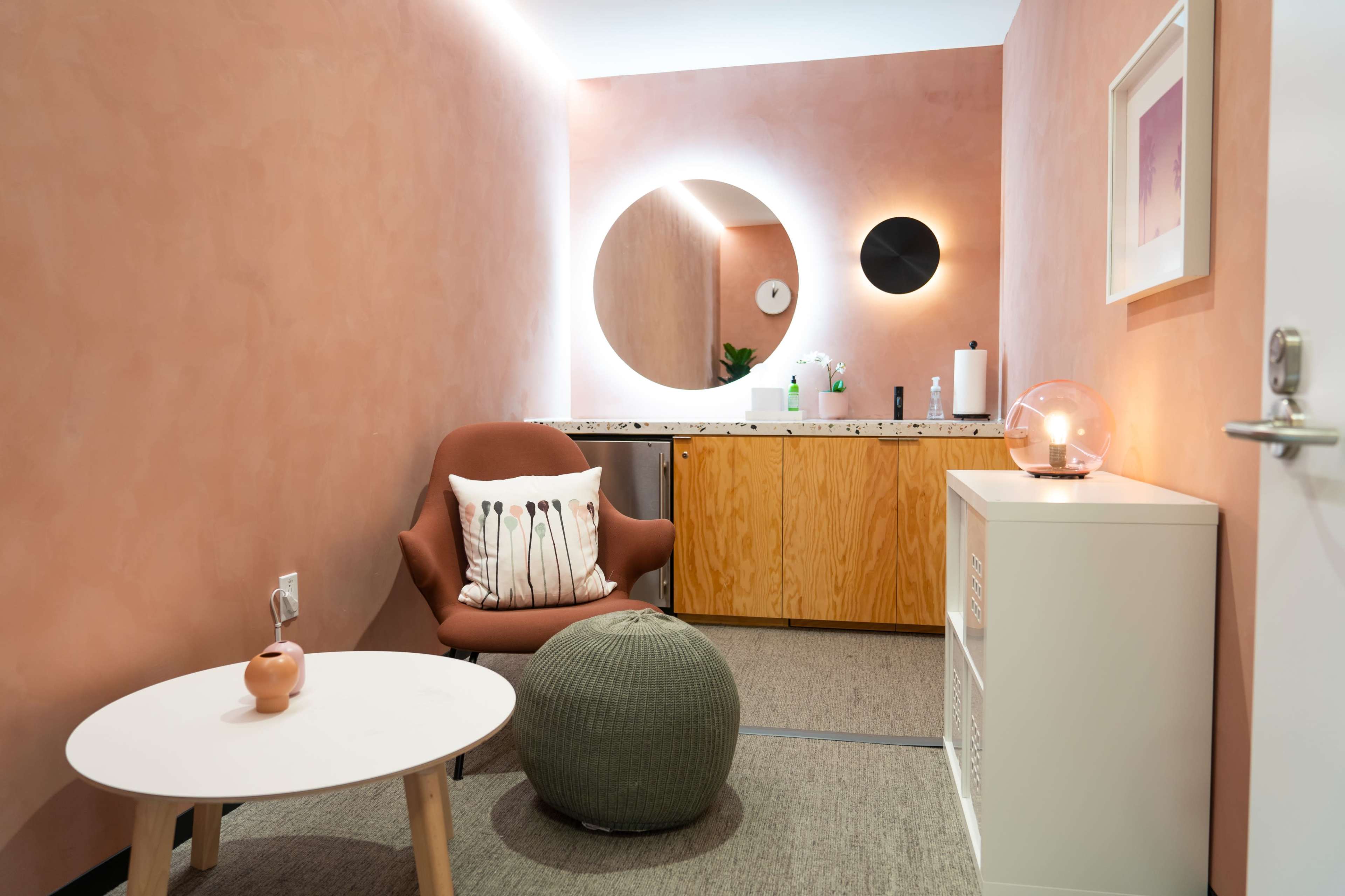 A cozy interior space featuring a round mirror with built-in lighting, a brown armchair, a small table, a green pouf, and a wooden cabinet against the softly colored walls.