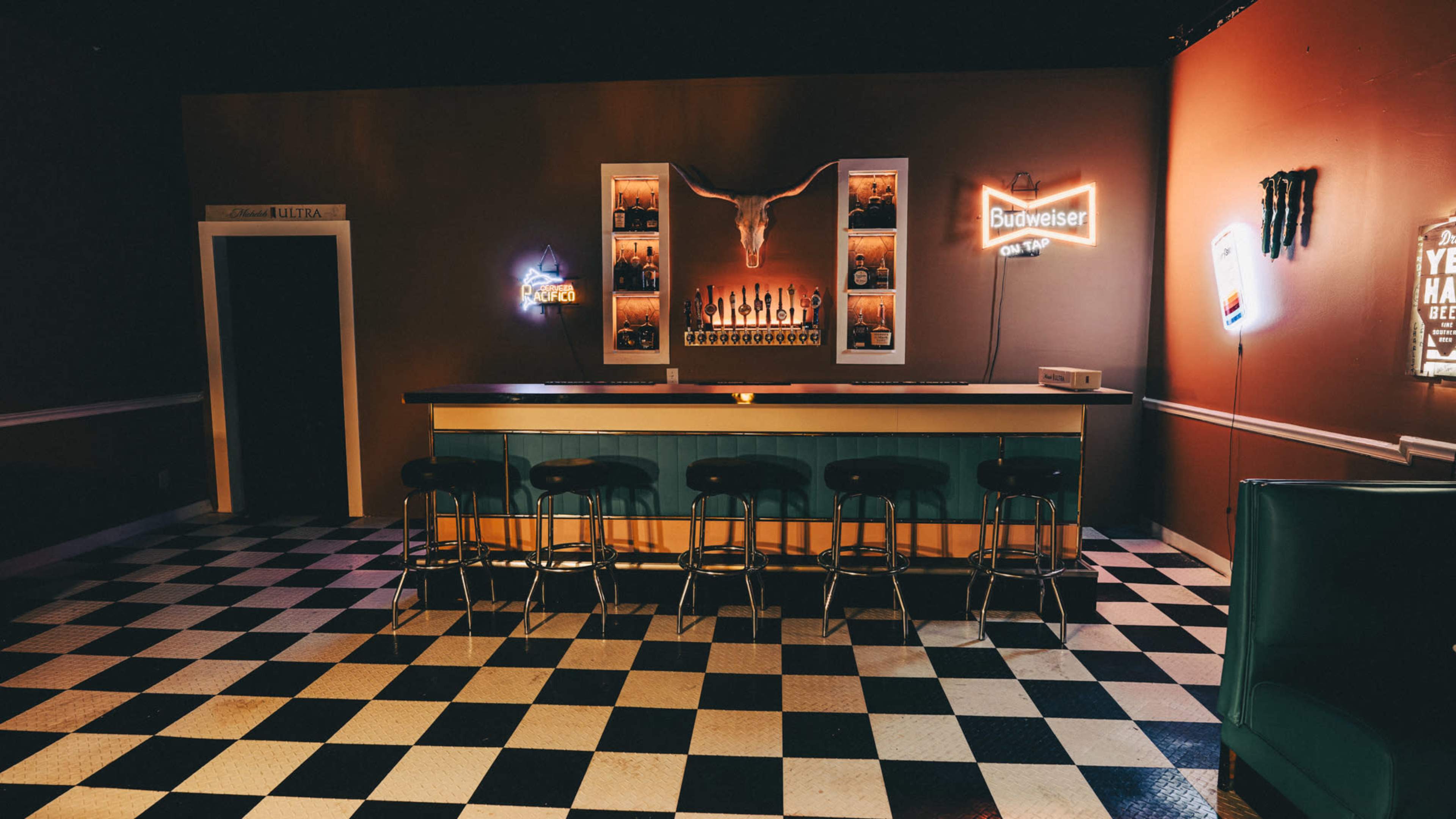 The image shows a dimly lit bar area with a black and white checkered floor, a long bar counter adorned with bottles and a decorative bull skull on the wall.