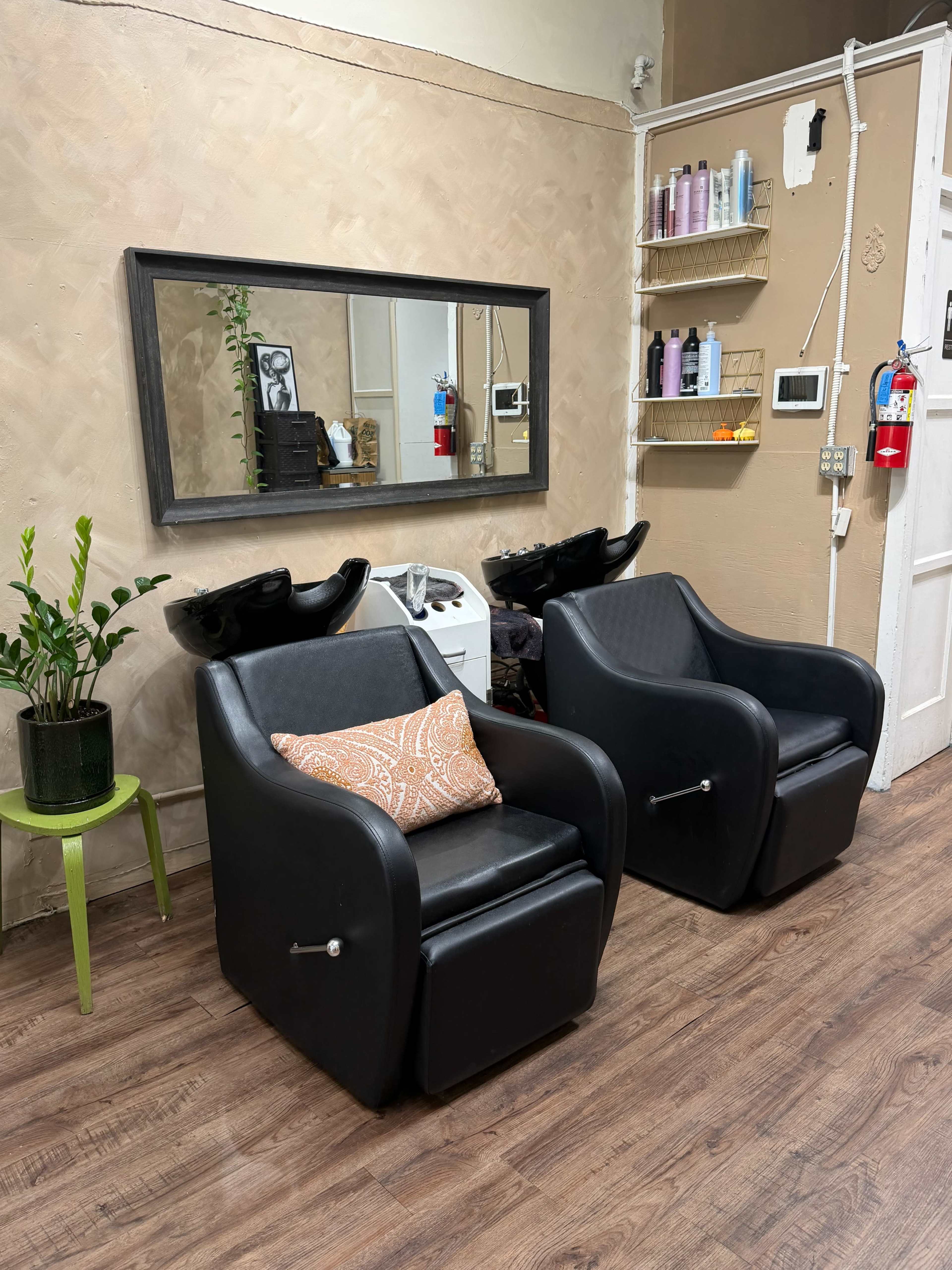 The image shows a hair salon interior featuring two black salon chairs with a decorative pillow, a large mirror on the wall, and hair care products displayed on shelves.