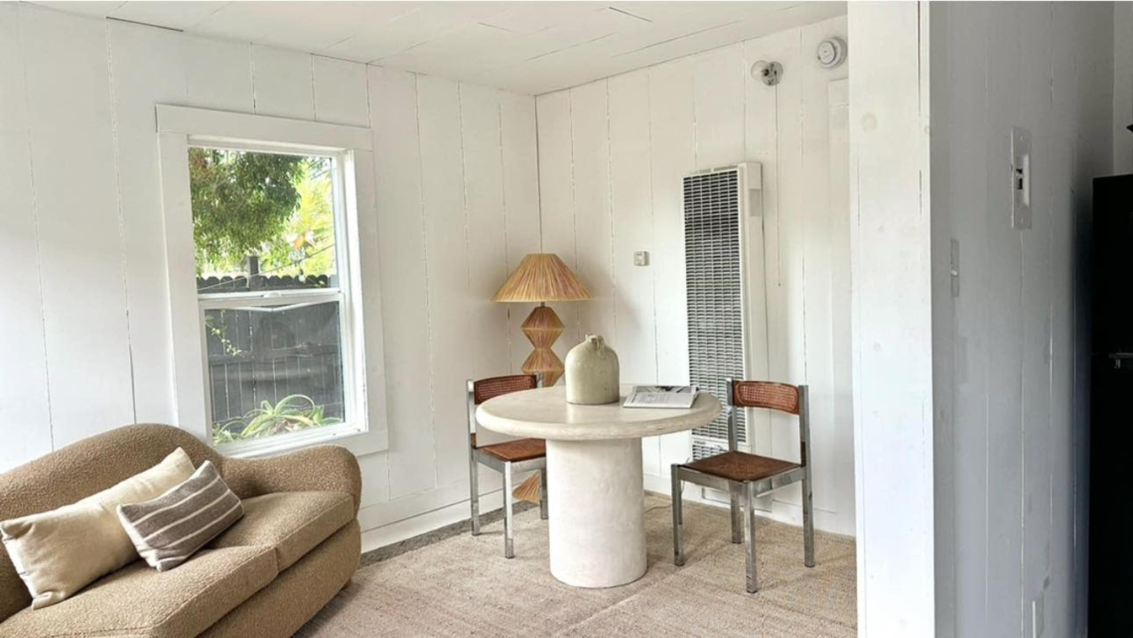 The scene features a small, minimalist dining area with a round table, two wooden chairs, and a sofa, all set against a backdrop of white walls and a window.