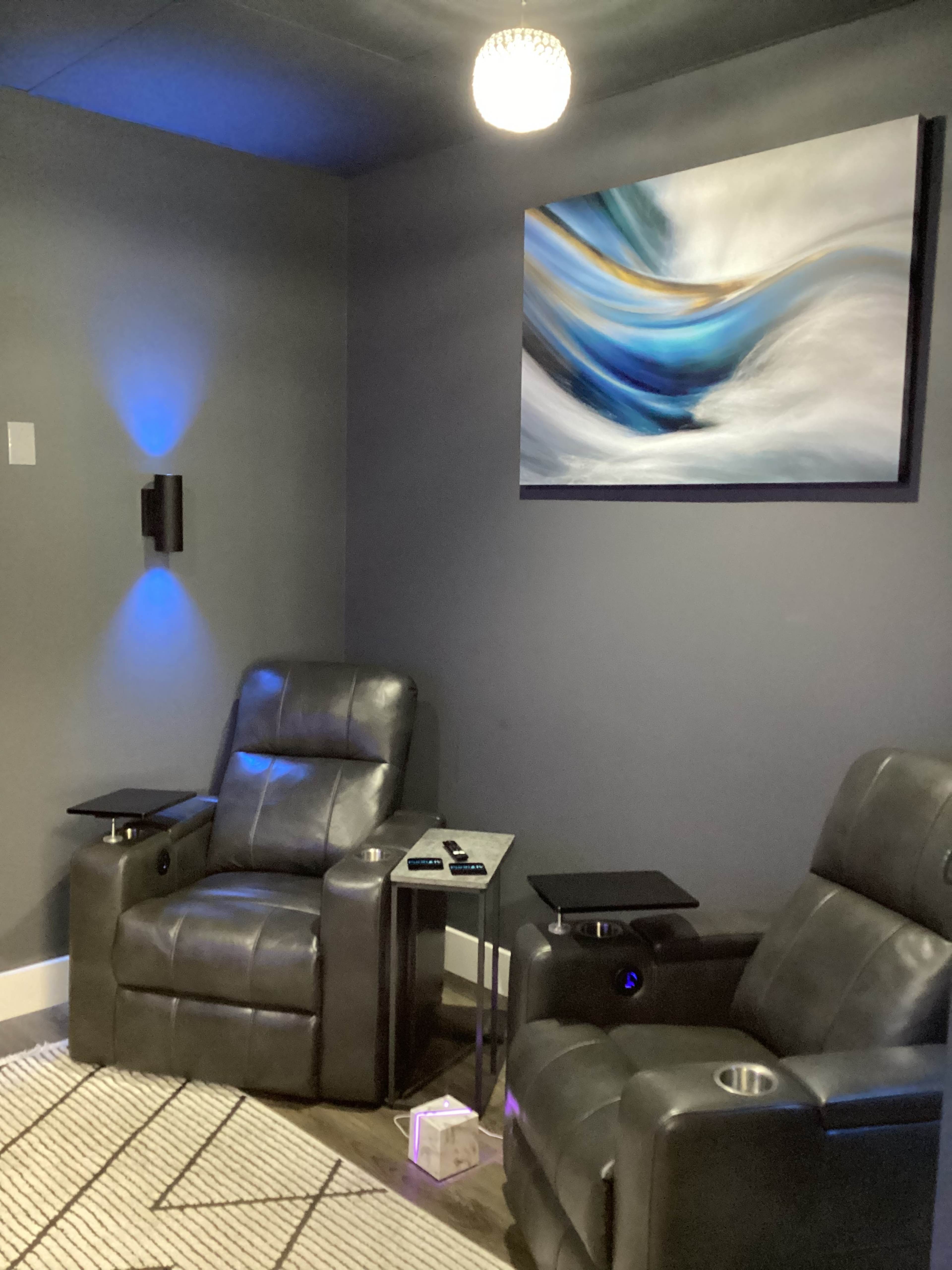 A small home theater setup with two gray recliner chairs, a table between them, and a colorful abstract painting on the wall.