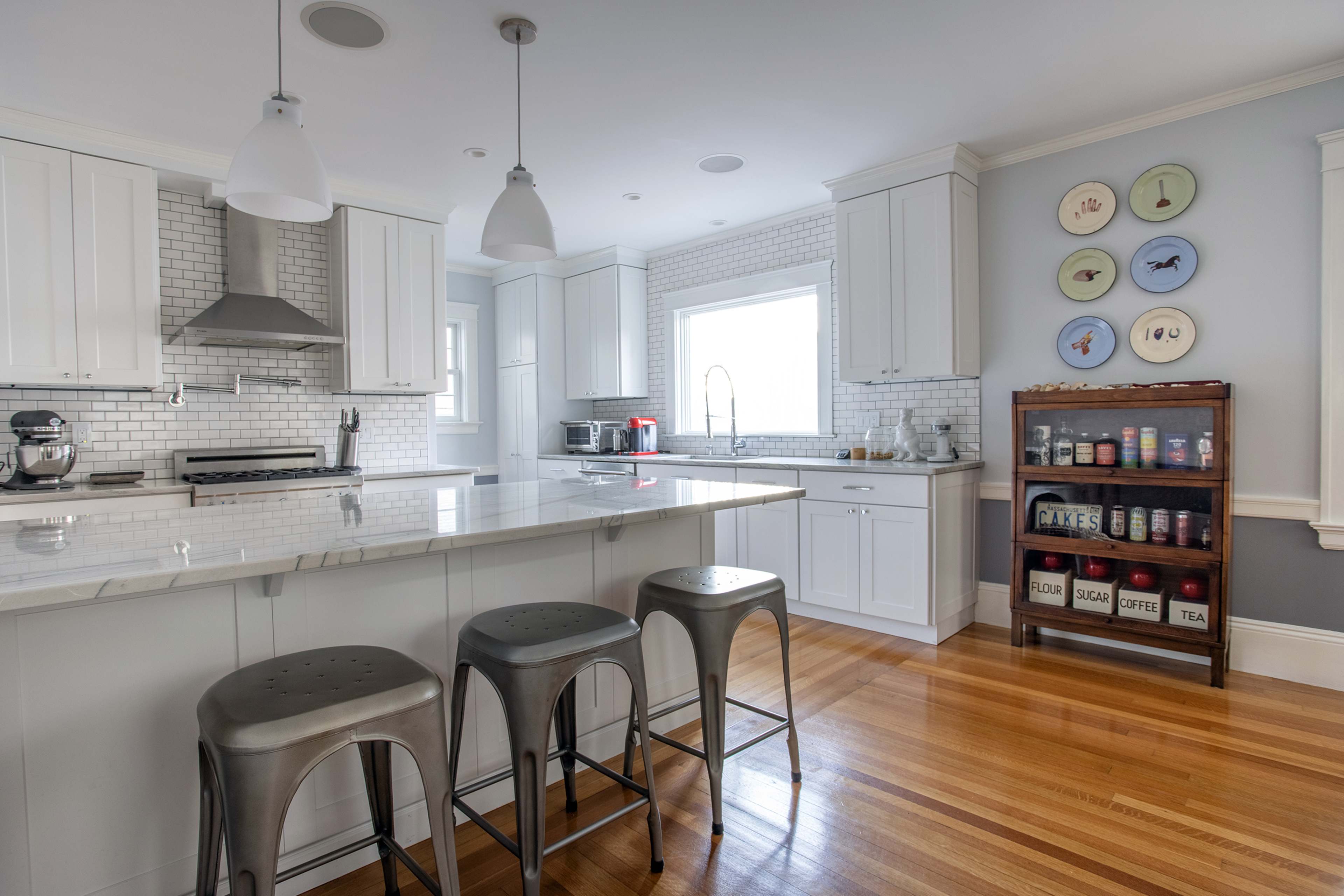 The kitchen features white cabinetry, a marble countertop island with three metal stools, and a wall of backsplash tiles.