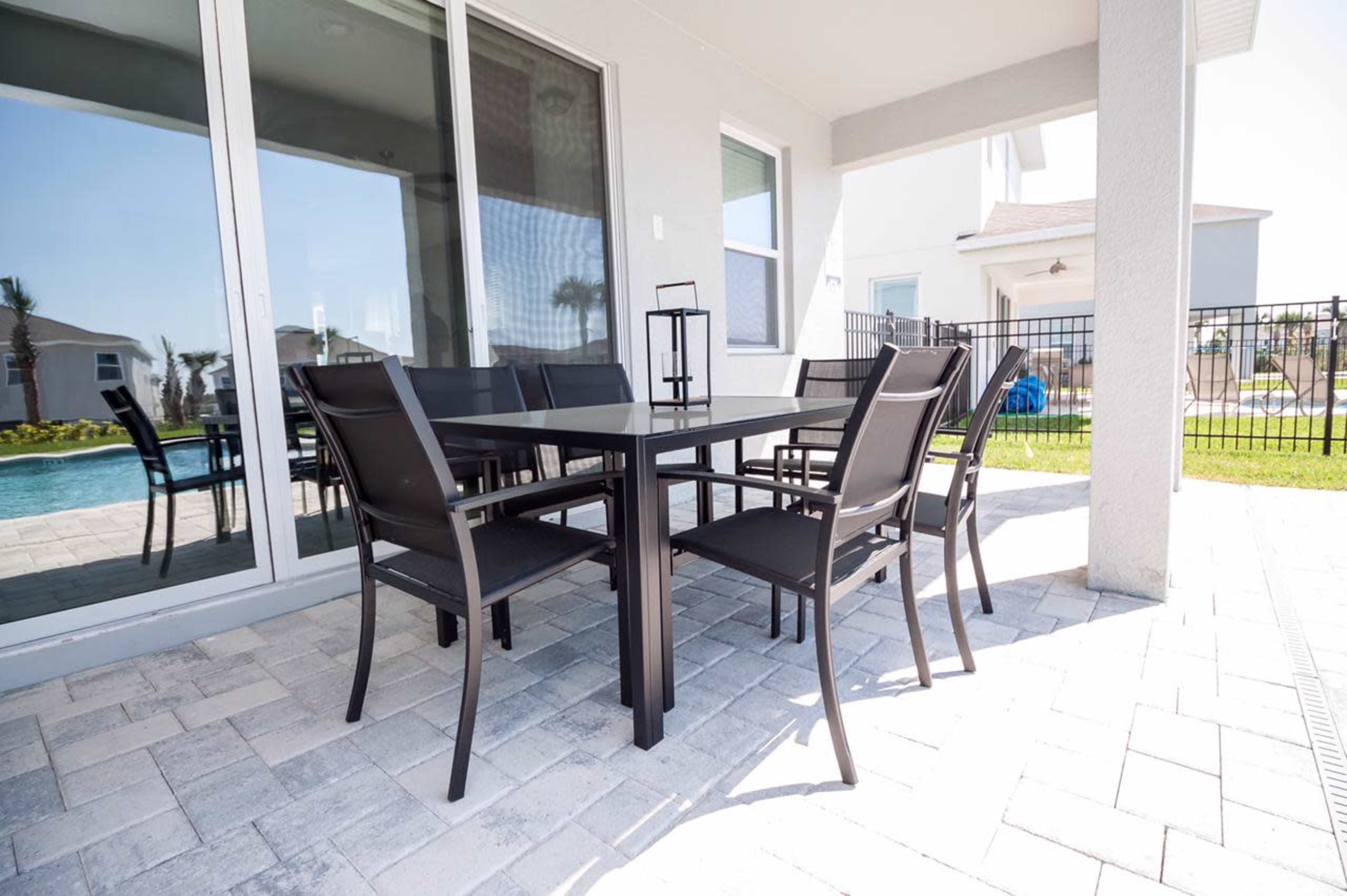 A patio area features a rectangular table surrounded by six chairs, with sliding glass doors in the background and a pool visible beyond a fence.