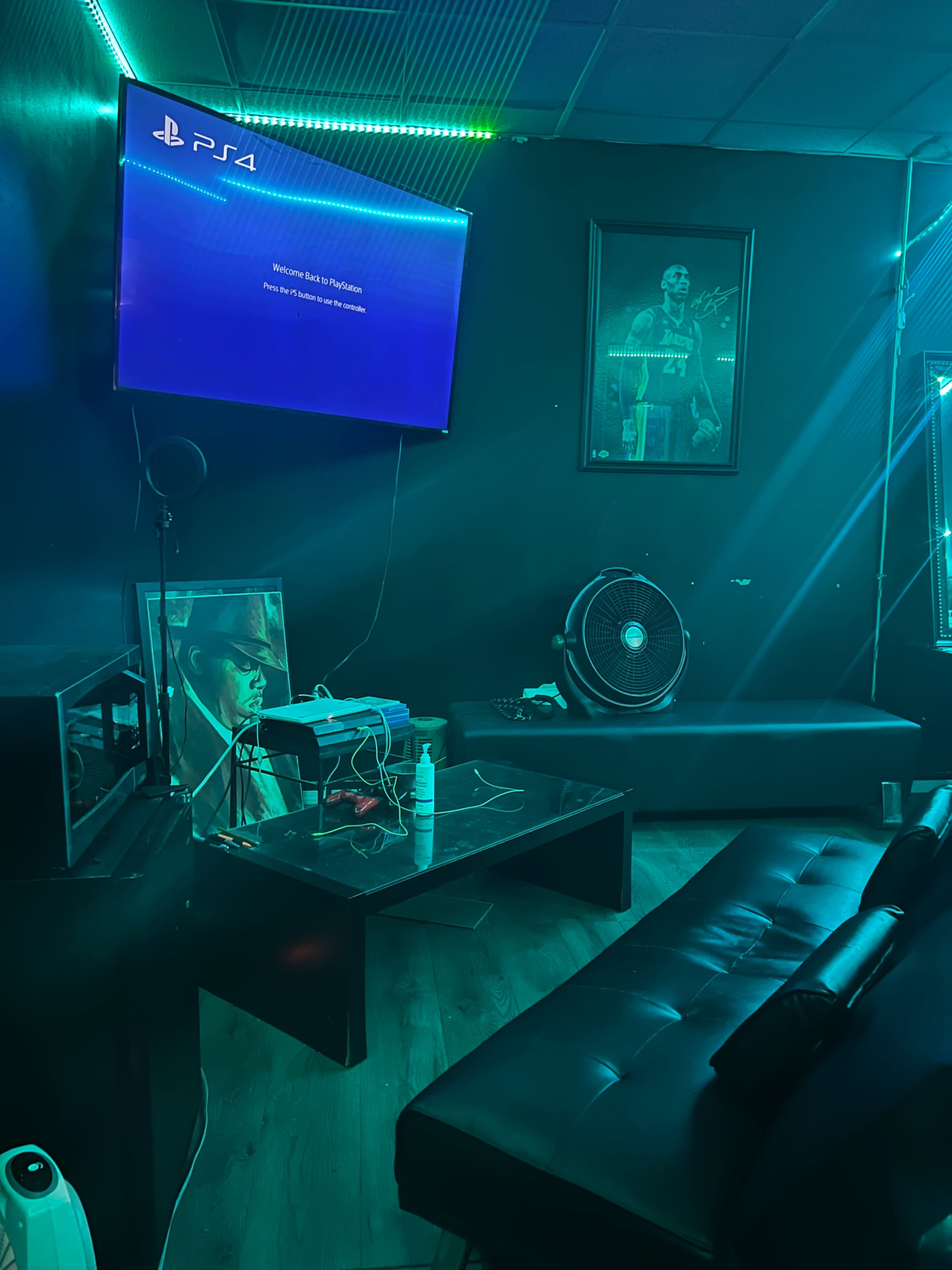 A gaming room features a PS4 console displayed on a wall-mounted screen, with a dark color scheme and furniture including a leather couch and a table.