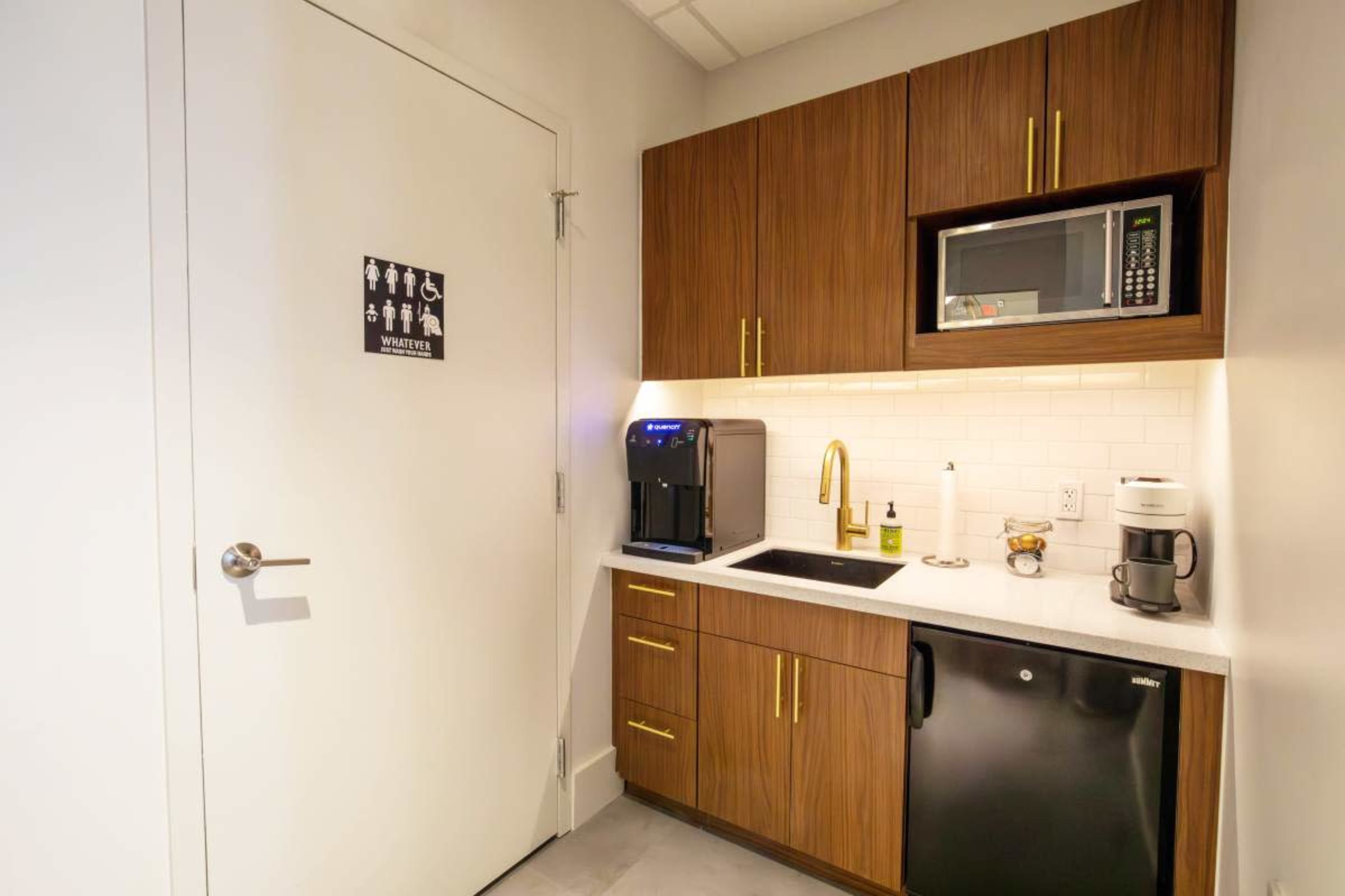 The image shows a small kitchenette featuring dark wooden cabinets, a sink, a coffee maker, a microwave, and a refrigerator, with a sign on the door indicating a restroom.