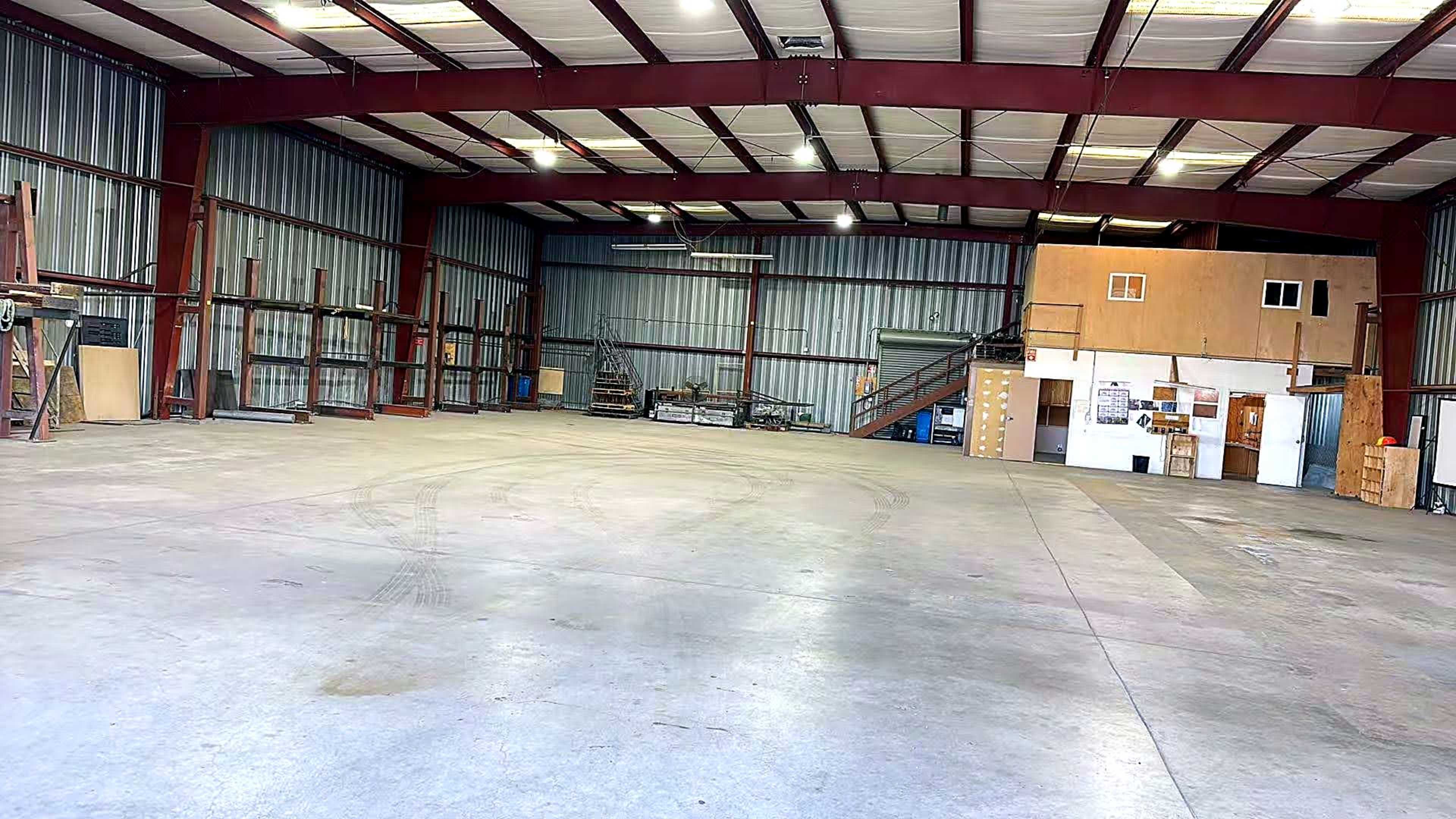 The image shows a spacious industrial warehouse with high ceilings, metal beams, and concrete flooring, featuring various storage materials along the walls.