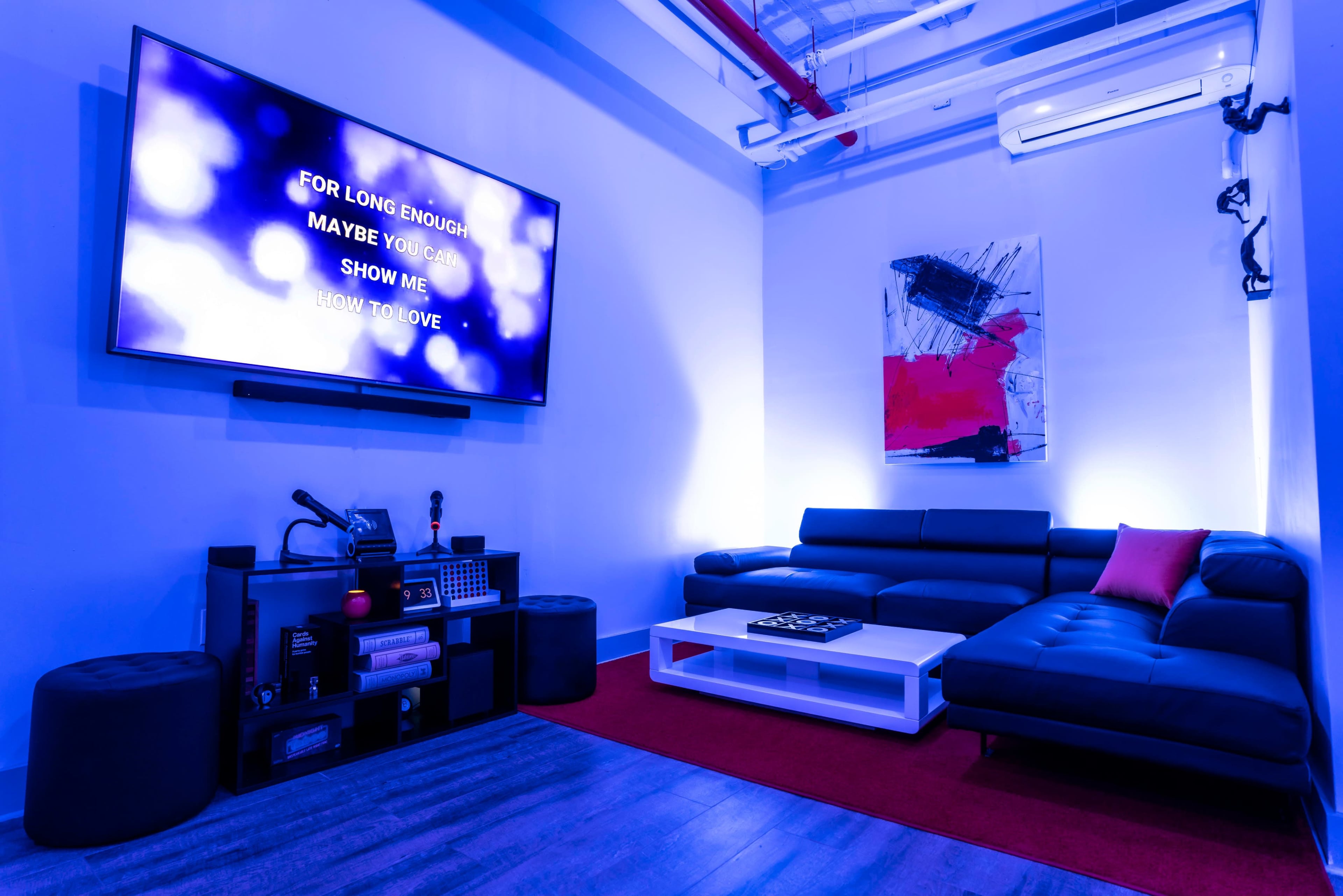 The image shows a modern lounge area with a large wall-mounted television displaying lyrics, a black sectional sofa, and a coffee table on a red carpet.