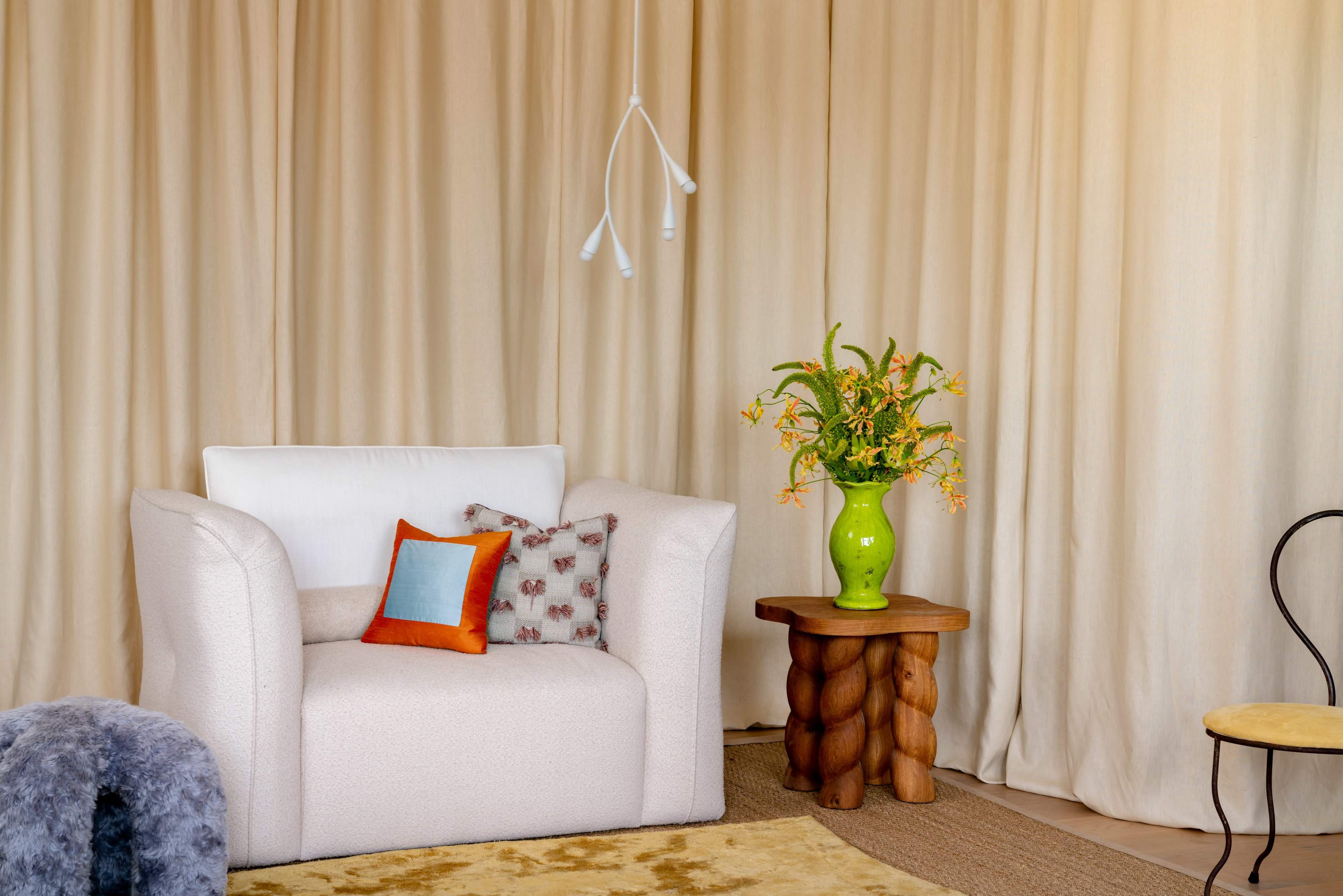 A cozy seating area features a white armchair with decorative cushions beside a wooden side table adorned with a green vase filled with flowers.
