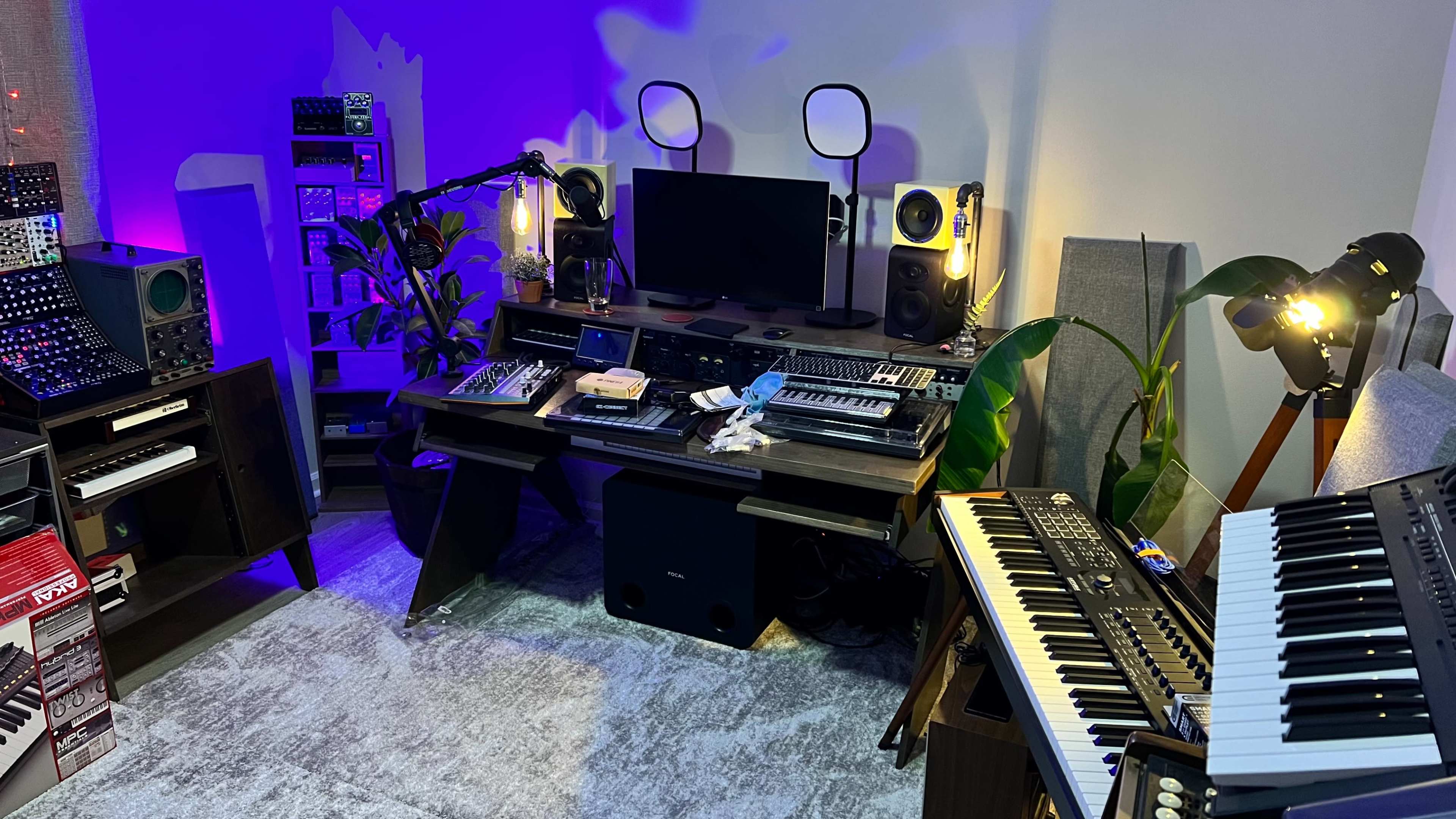 A music production studio with synthesizers, a computer monitor, and various audio equipment arranged around a central desk, with plants and ambient lighting in the background.