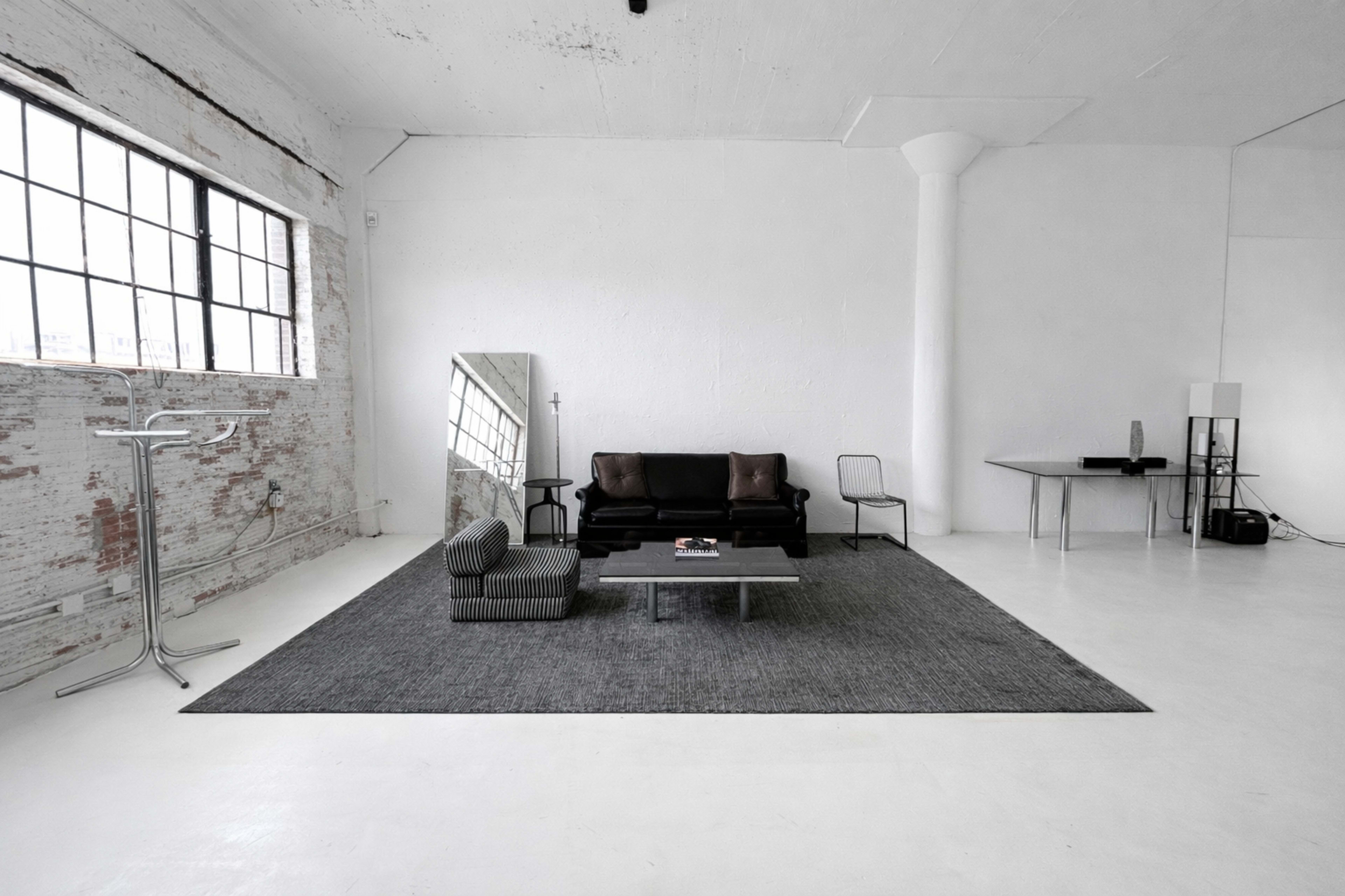 The image shows a minimalist interior space with a black sofa, a striped chair, a coffee table on a large gray rug, and large windows letting in natural light.