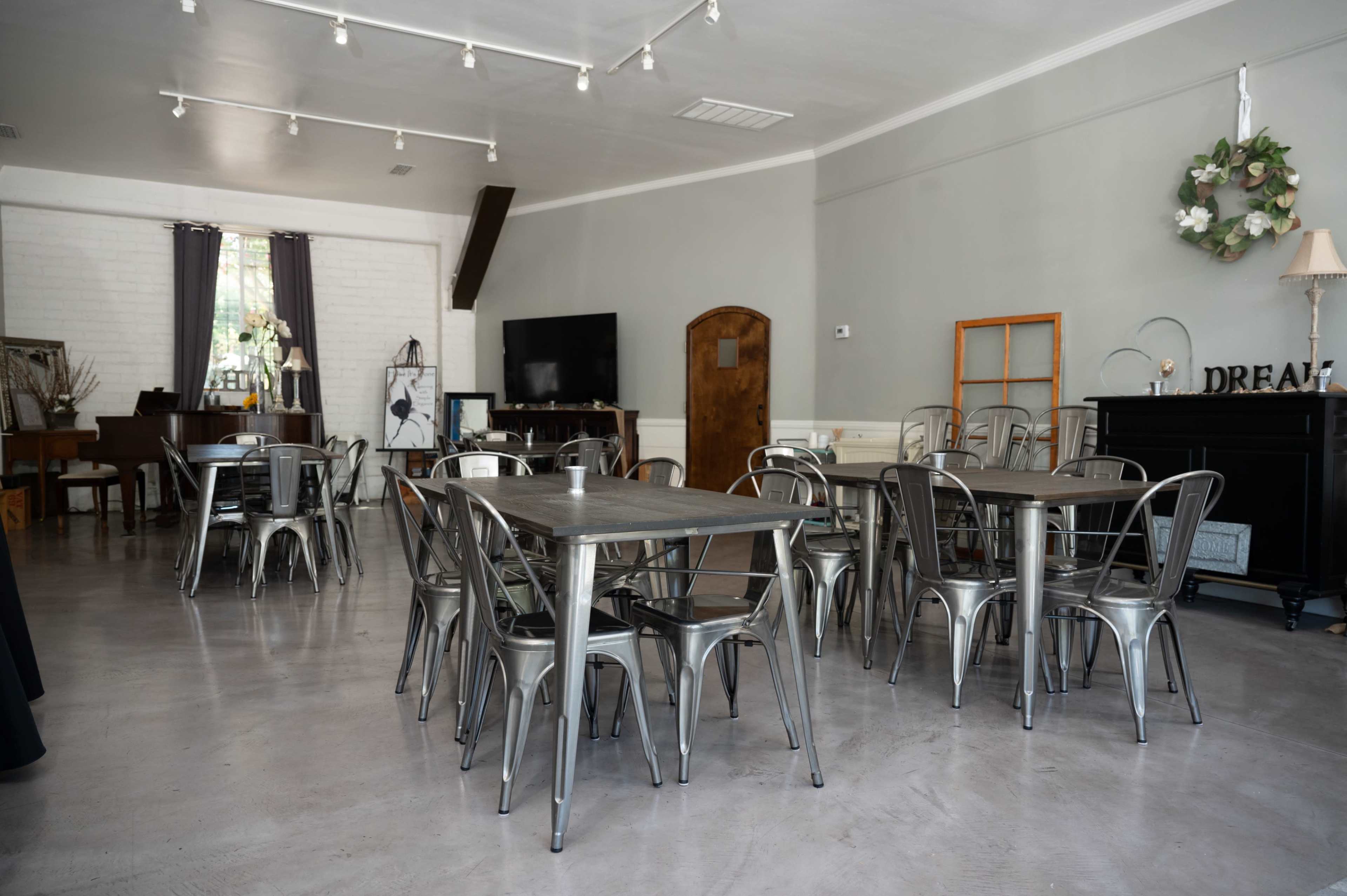 The image shows a spacious, modern room with metal tables and chairs arranged neatly, featuring a piano, a television, and a decorative window.