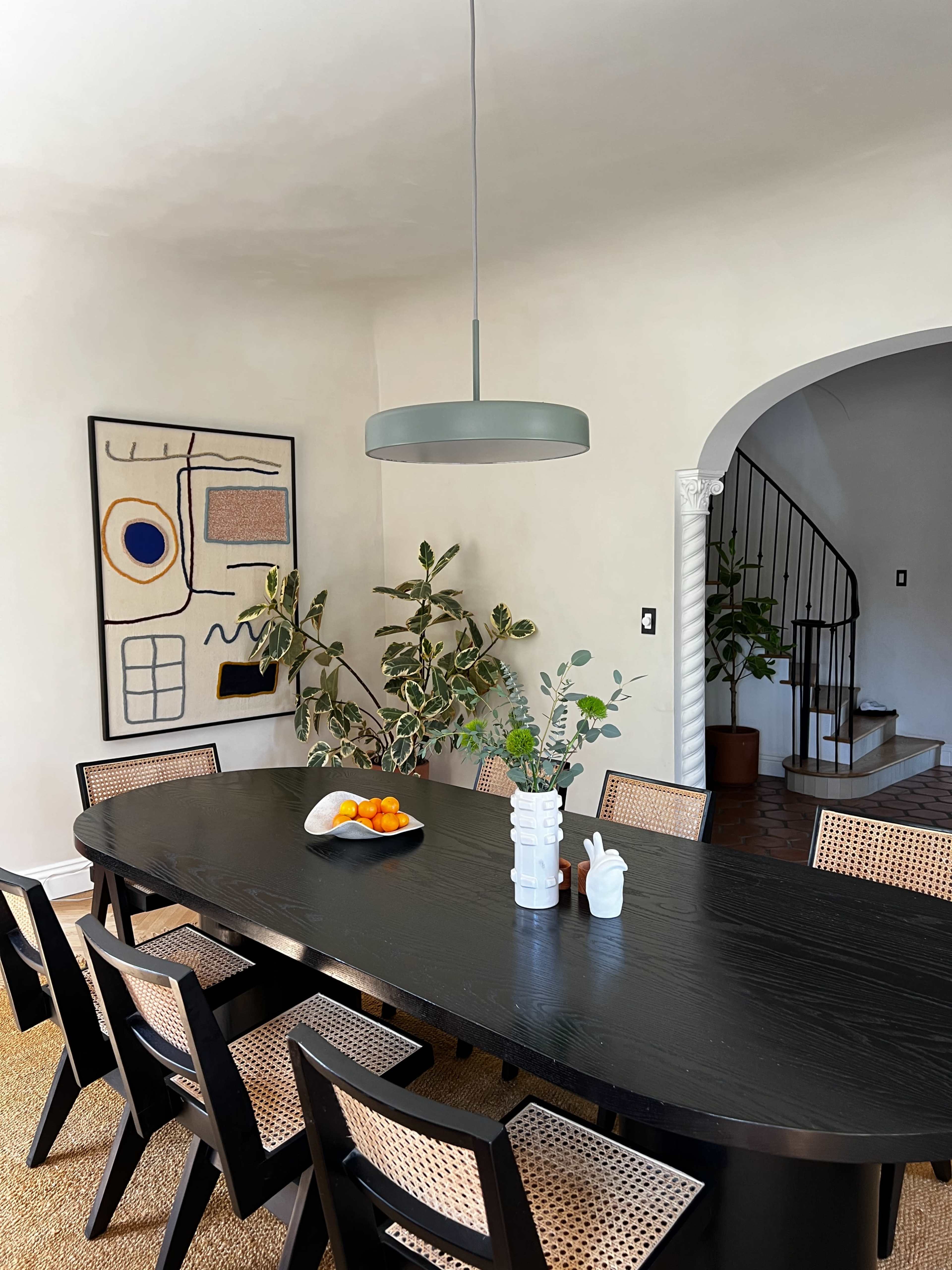 A large black dining table is set in a well-lit room, accompanied by chairs, a modern pendant light, a fruit bowl, and decorative plants.