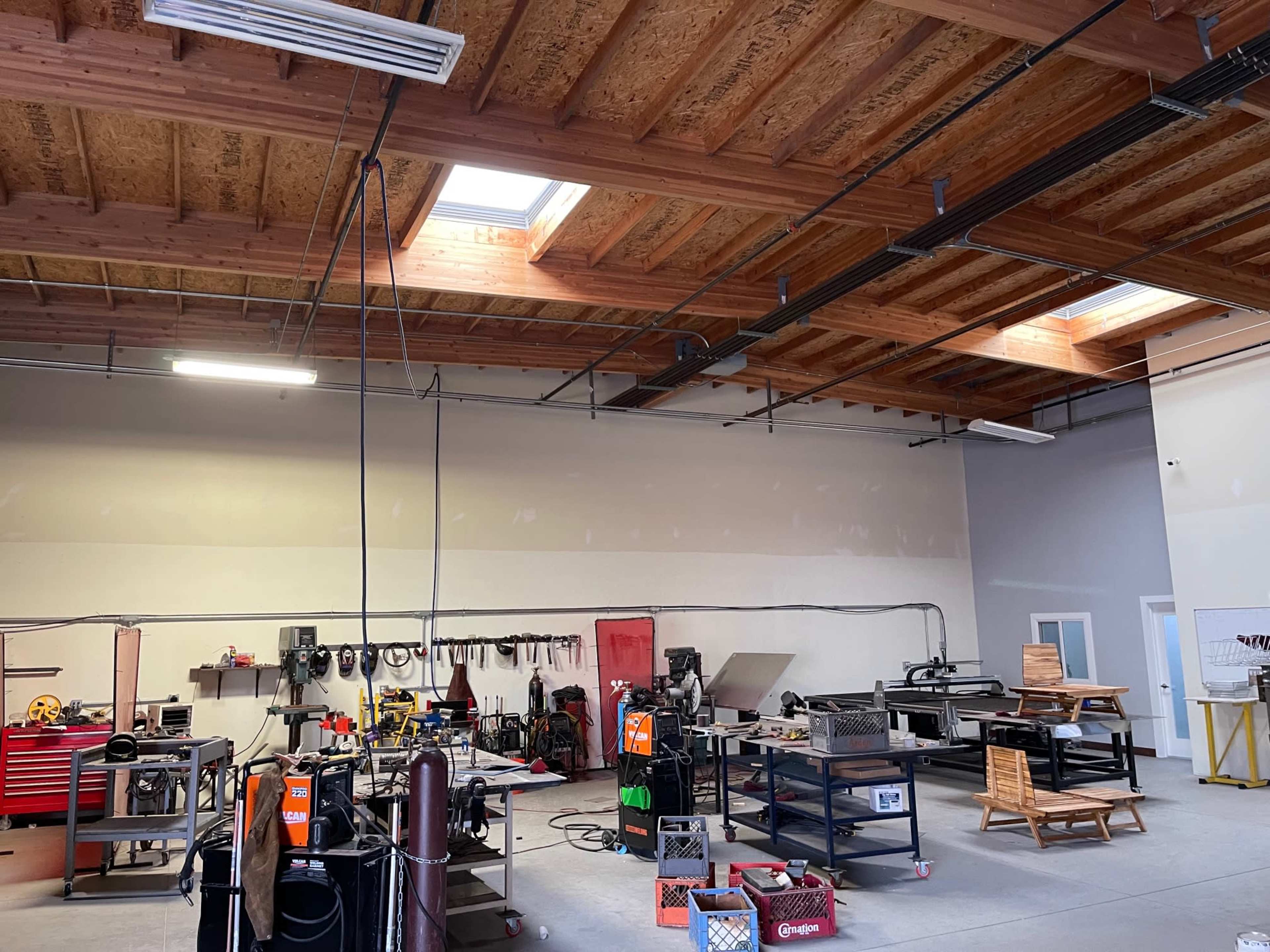 A spacious workshop featuring various tools and equipment, with skylights illuminating the area and workstations arranged along the walls.