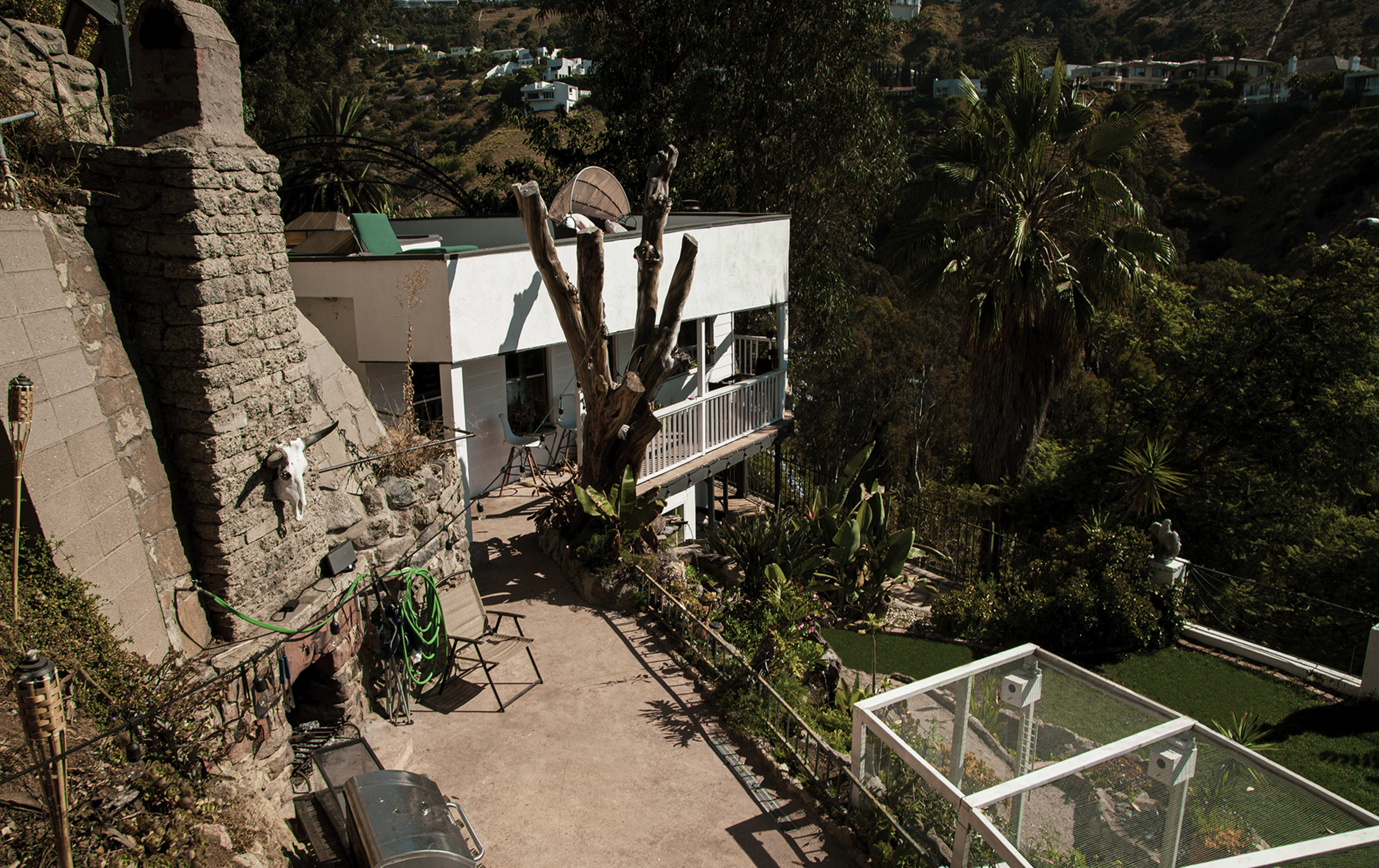 A hillside residence with a balcony, surrounded by greenery and a large tree, with a section of the yard featuring a glass enclosure.